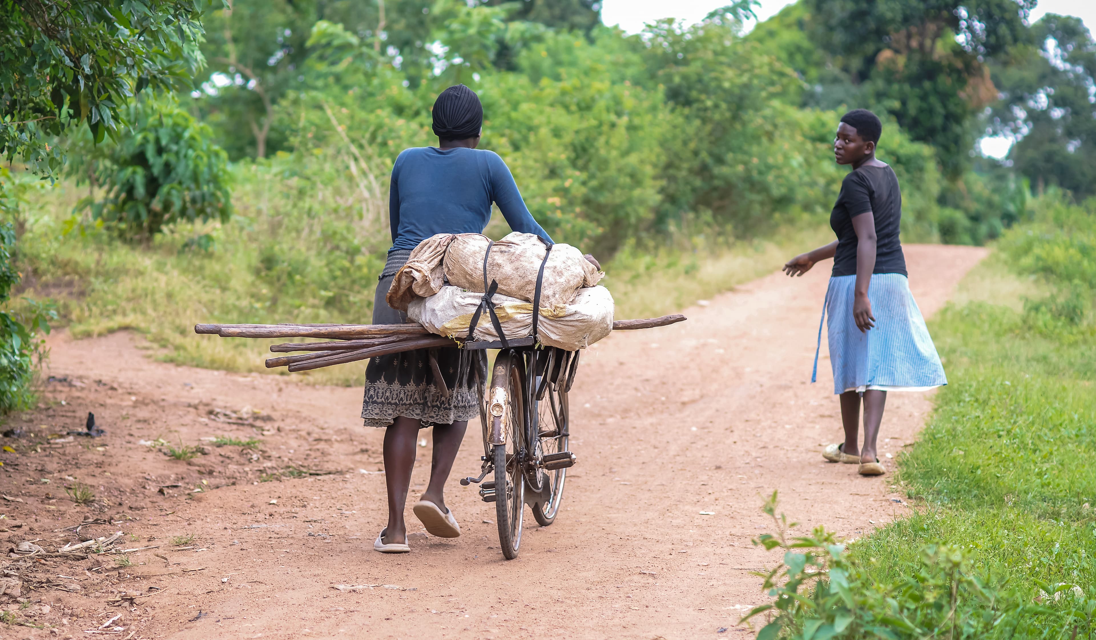 A man rides a bicycle loaded with jerry cans while a woman carrying a blue bag walks beside him on a dirt road.