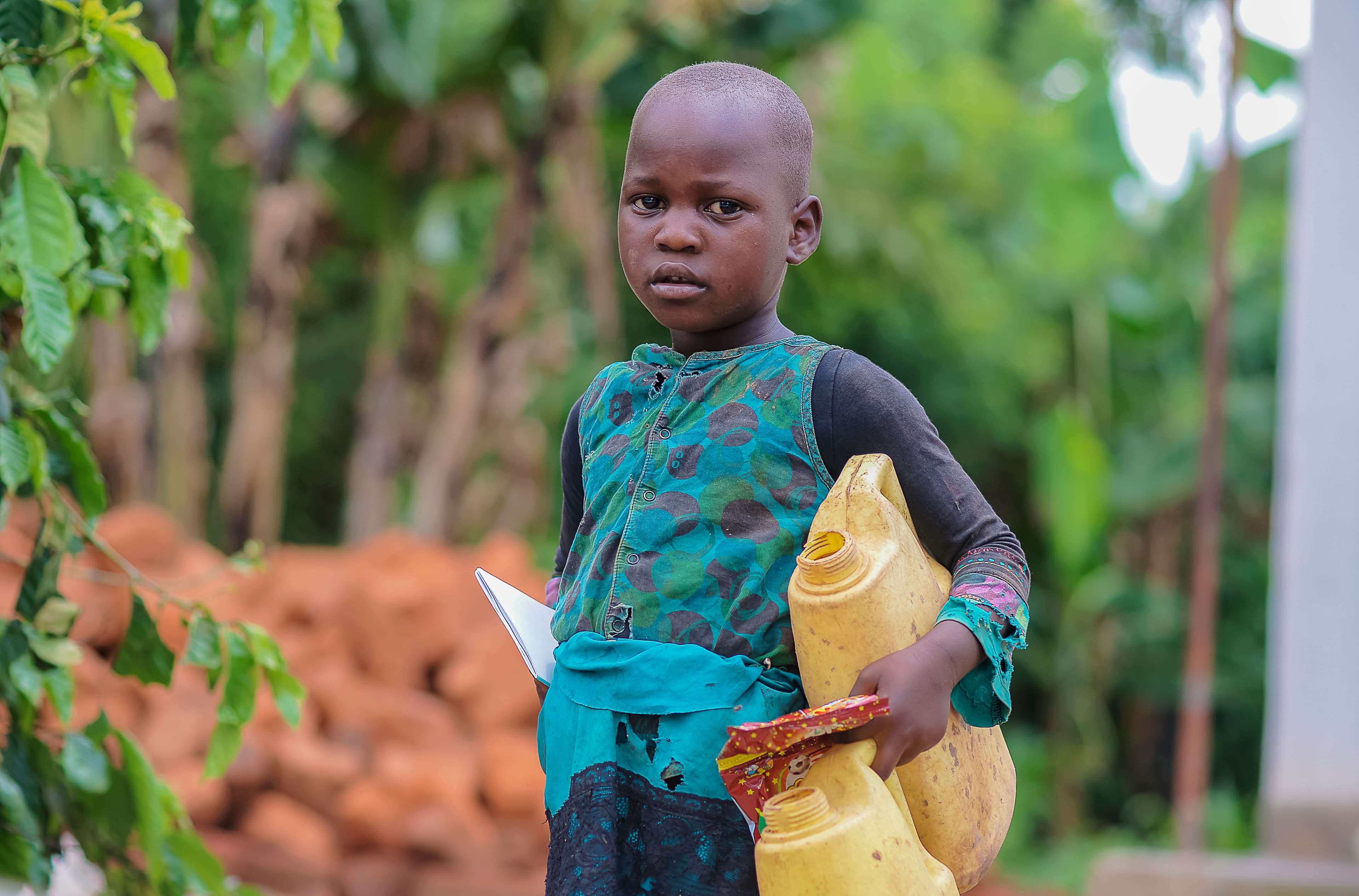 A boy in traditional dress stands with a yellow jerry can and a food container.