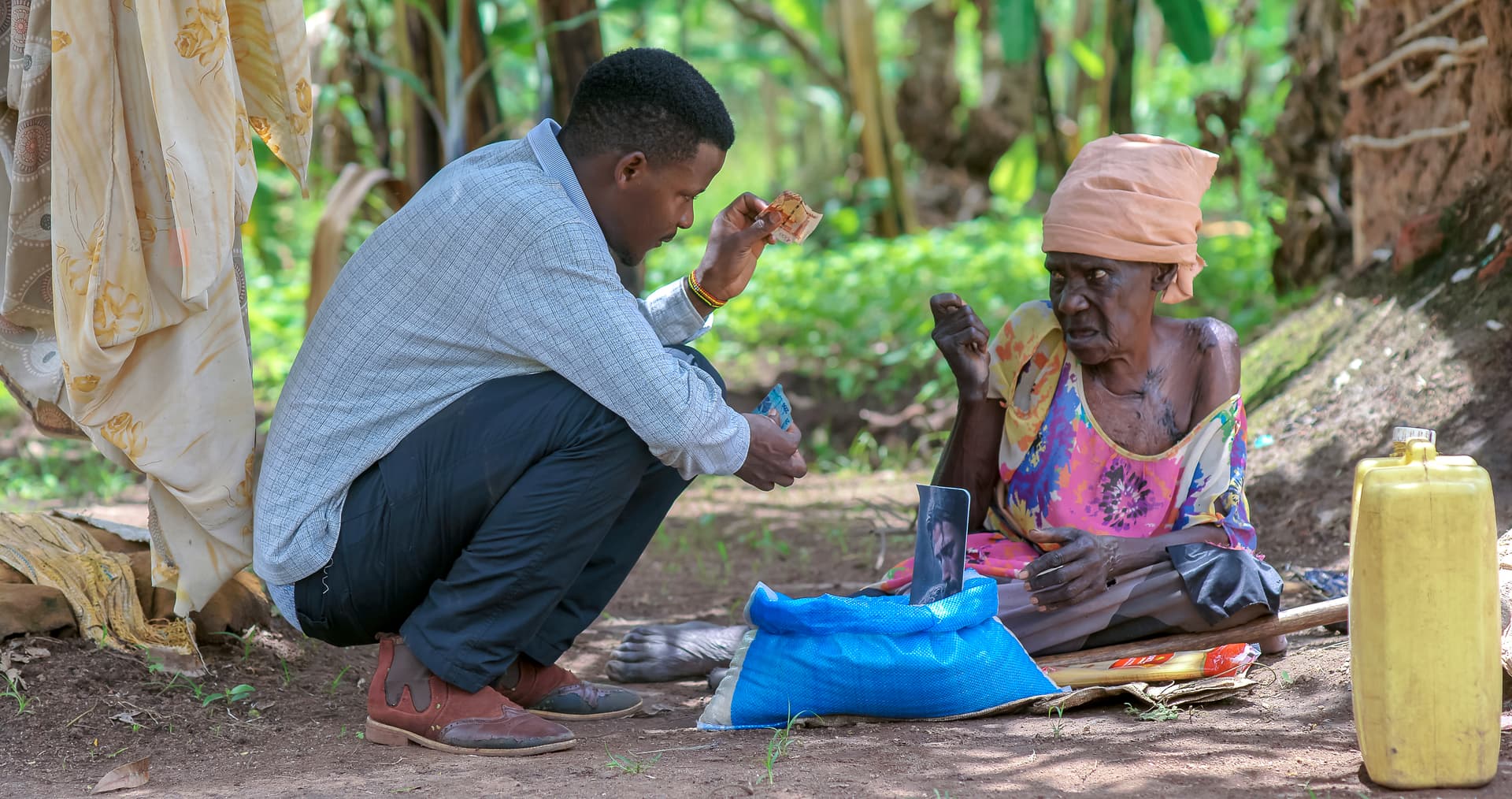 A JFM volunteer kneels beside Elder Atim Justine outside her hut, delivering a food gift package.