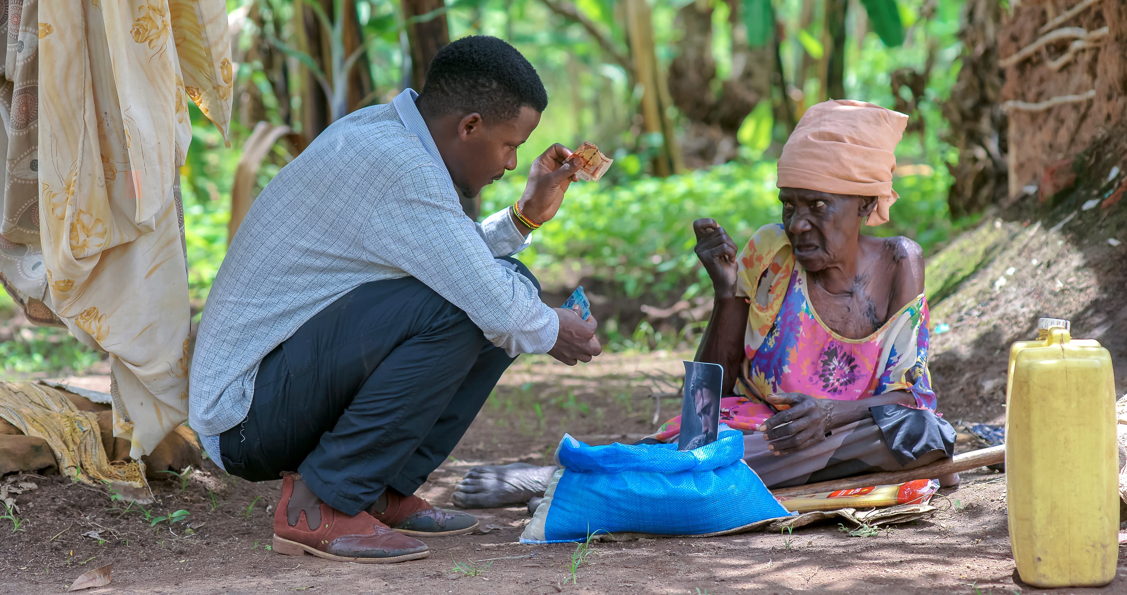 A JFM volunteer kneels beside Elder Atim Justine outside her hut, delivering a food gift package.