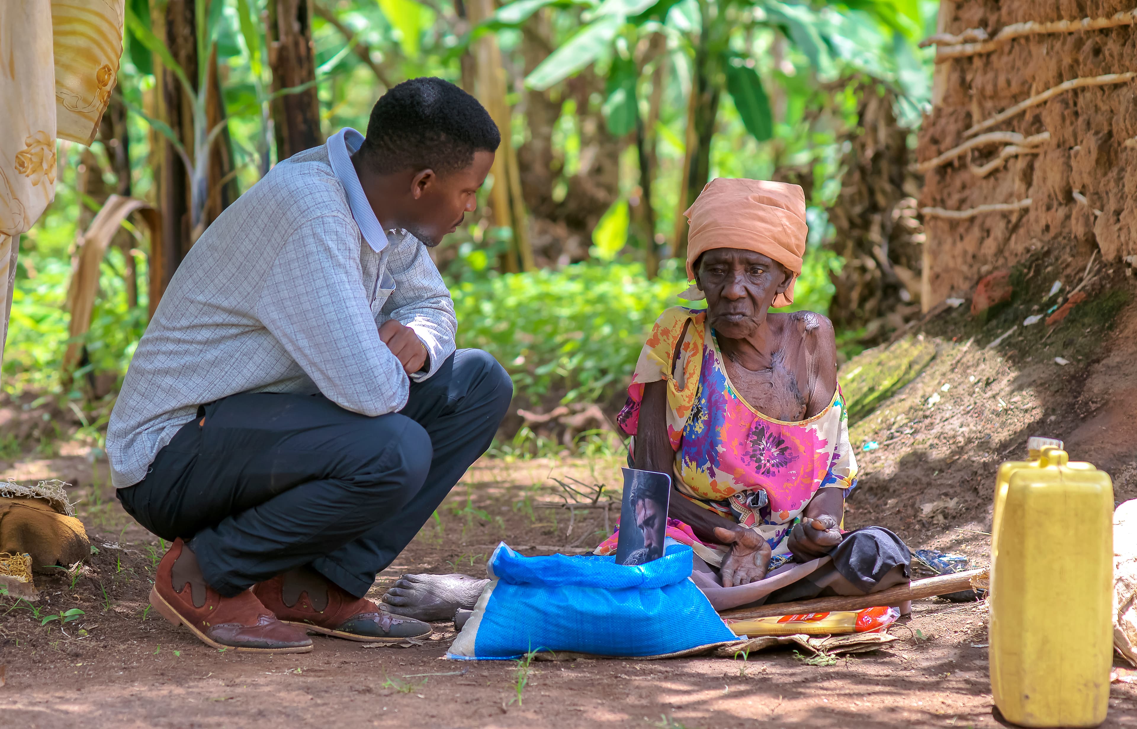 A JFM team member crouches beside elderly Elder Atim Justine, presenting her with a food gift package.