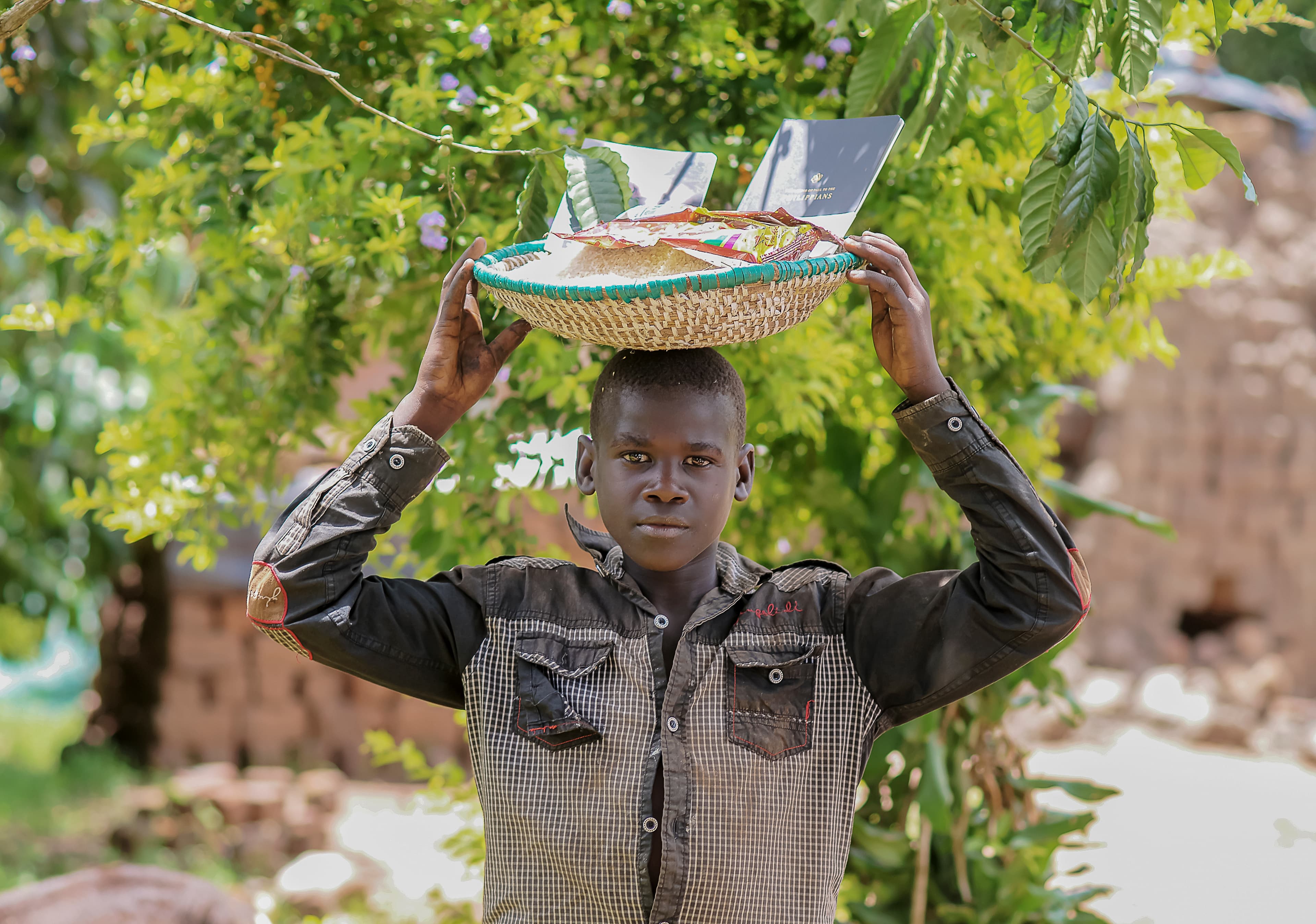 A boy balances a basket of food on his head near village buildings.