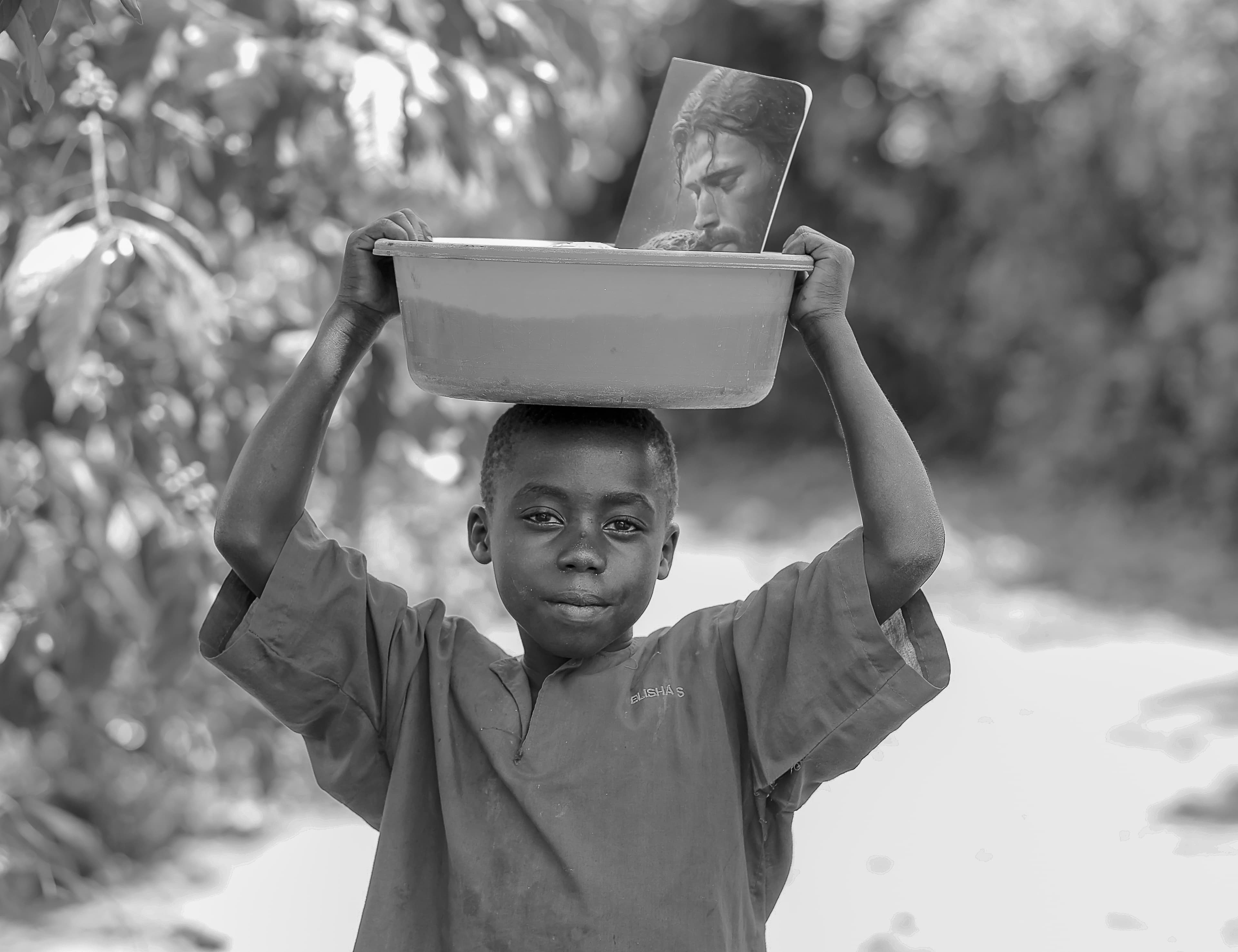 Black and white photo of a boy carrying a bowl on his head along a dirt path.
