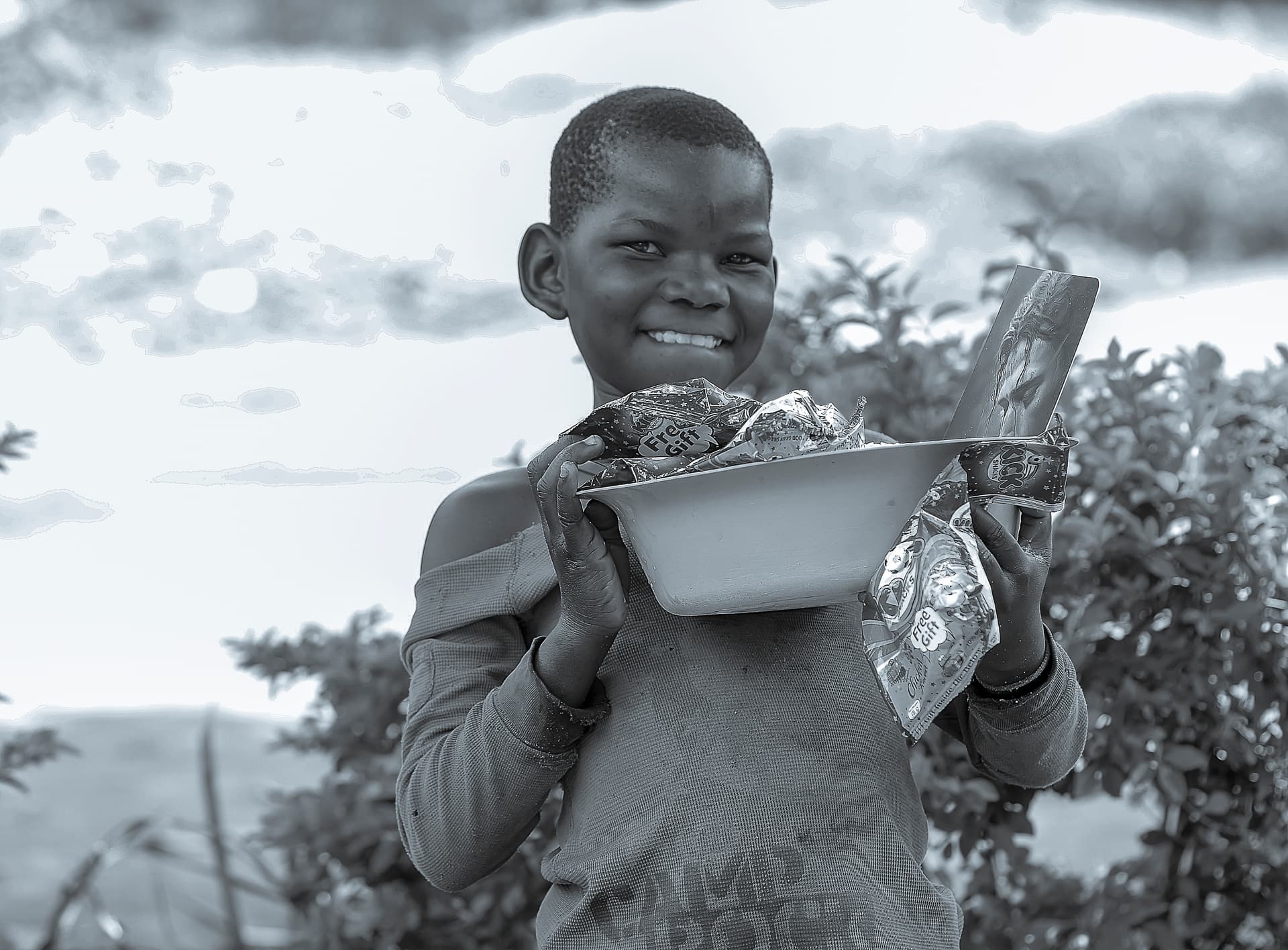 Black and white photo of a boy smiling while holding a bowl, standing near open water.