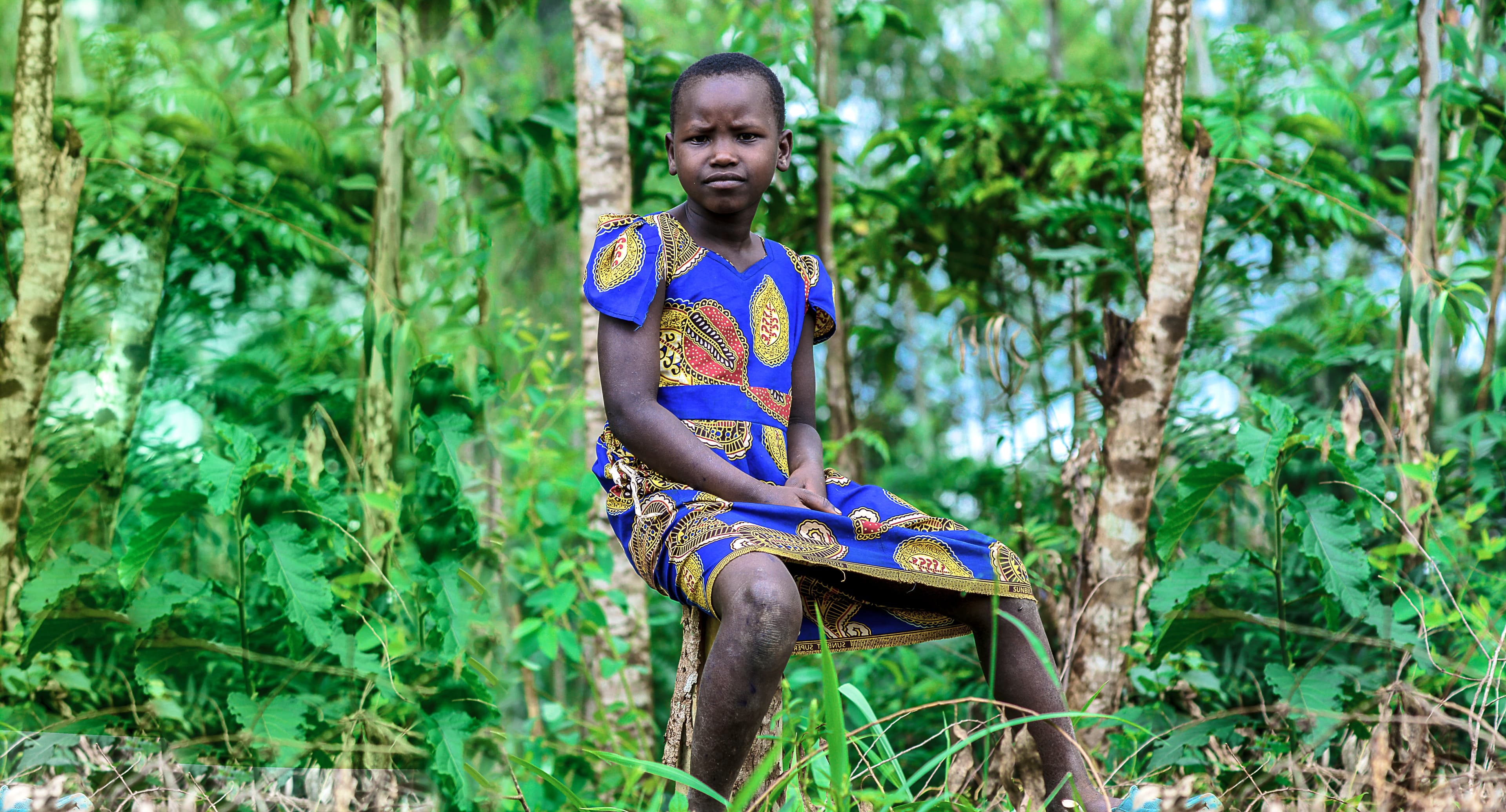 A girl in a patterned dress sits on a tree branch or log outdoors.