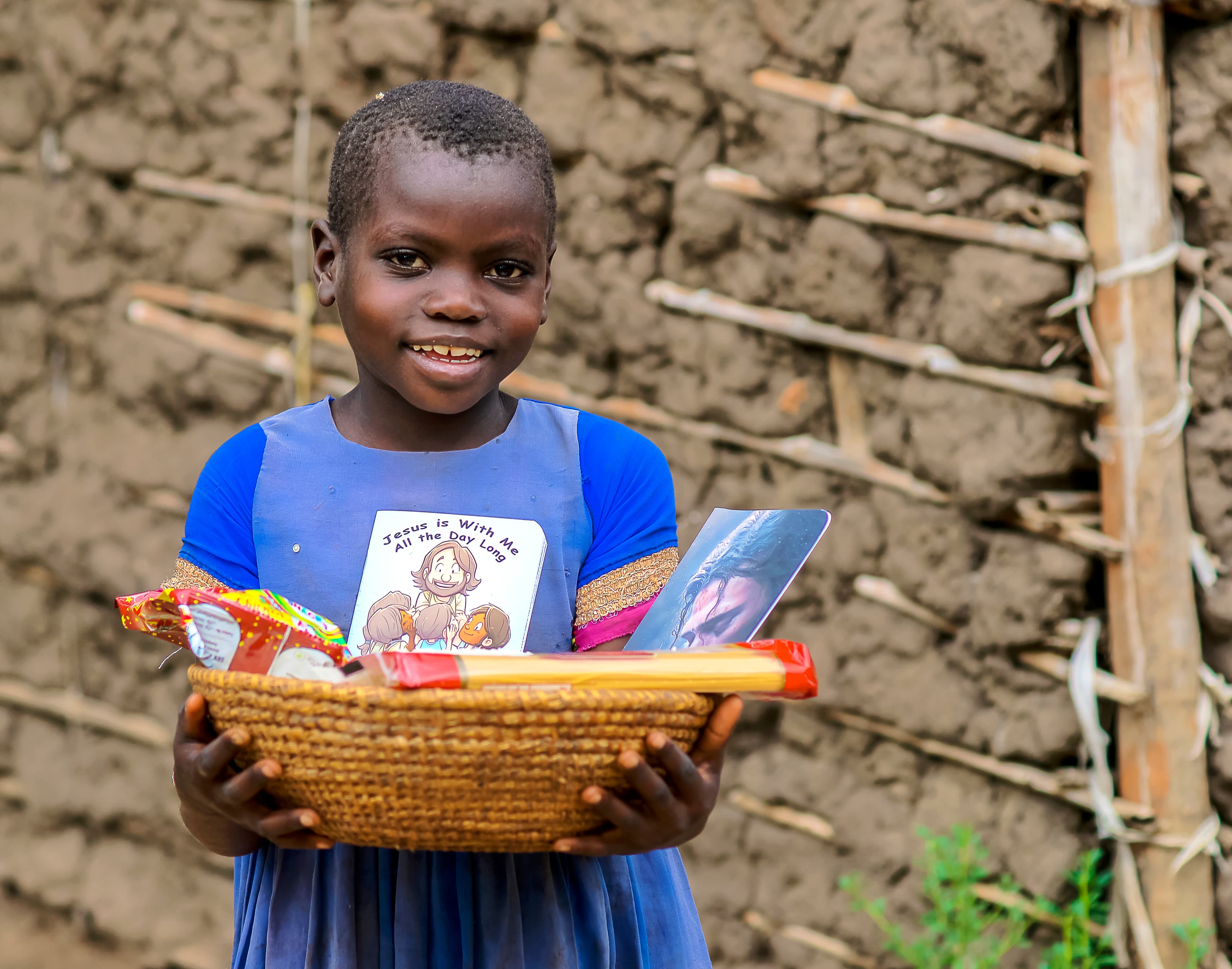 A girl stands near a mud house holding a basket of JFM food gift packages.