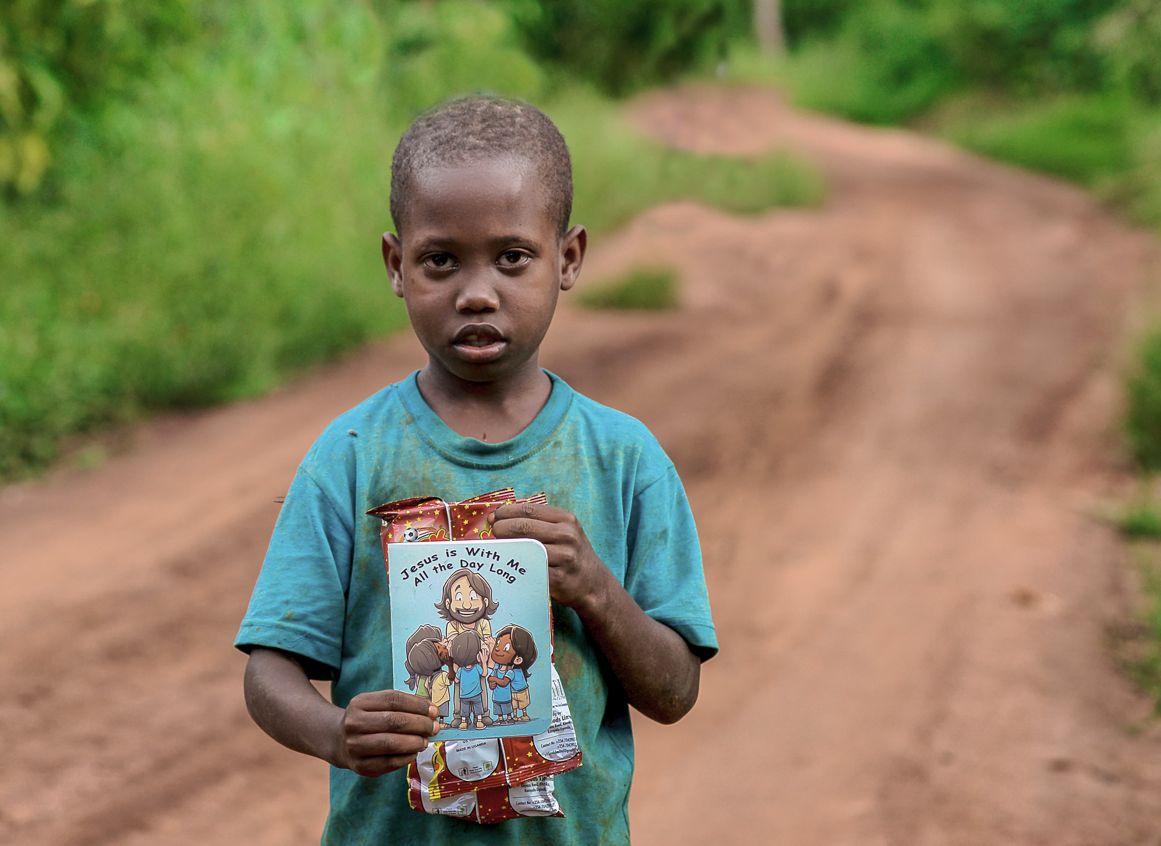 A boy stands on a dirt road holding a JFM food gift package and photo card.