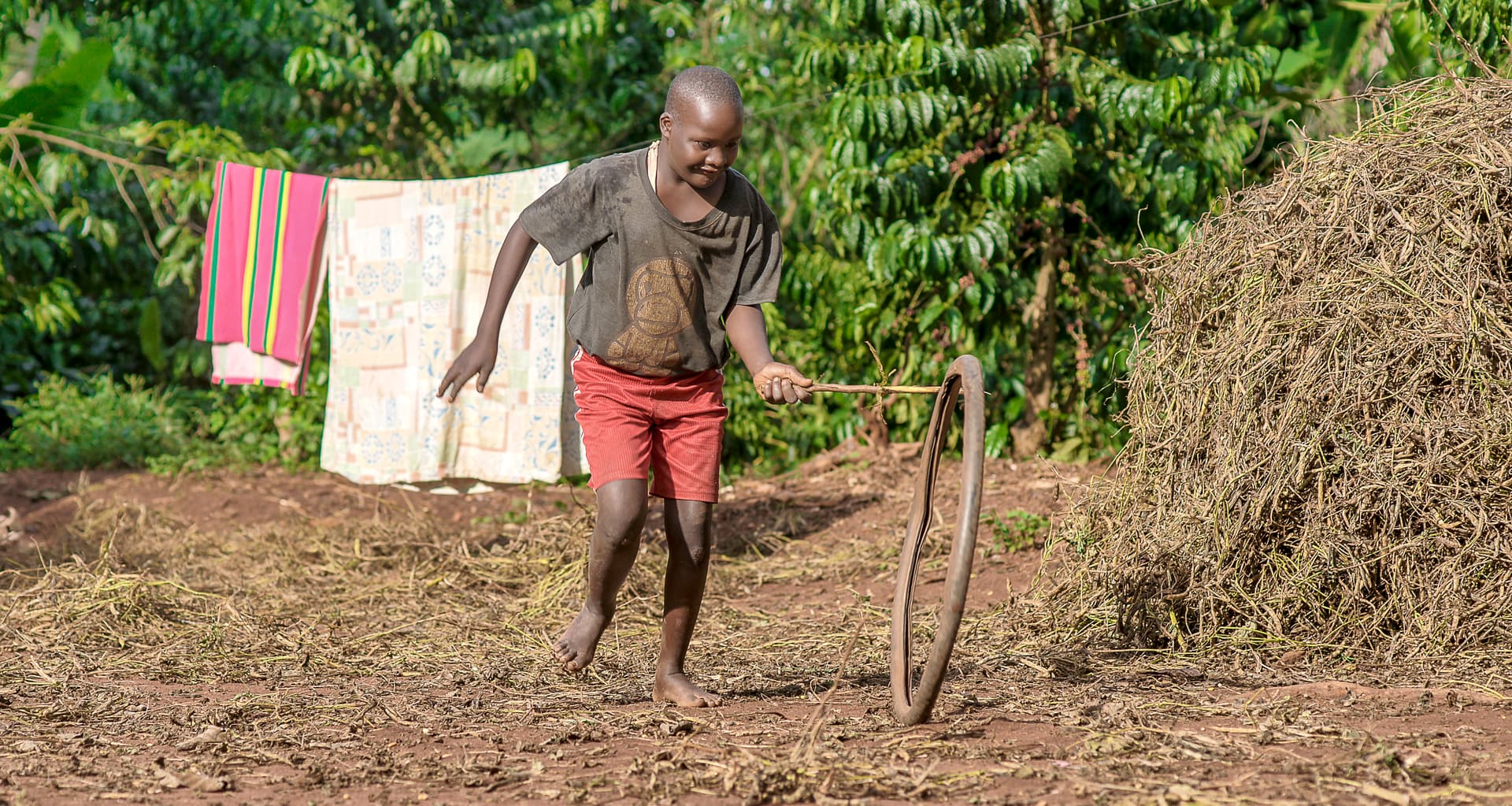 A shirtless boy runs outdoors past a clothesline of hanging laundry and a pile of dried grass.