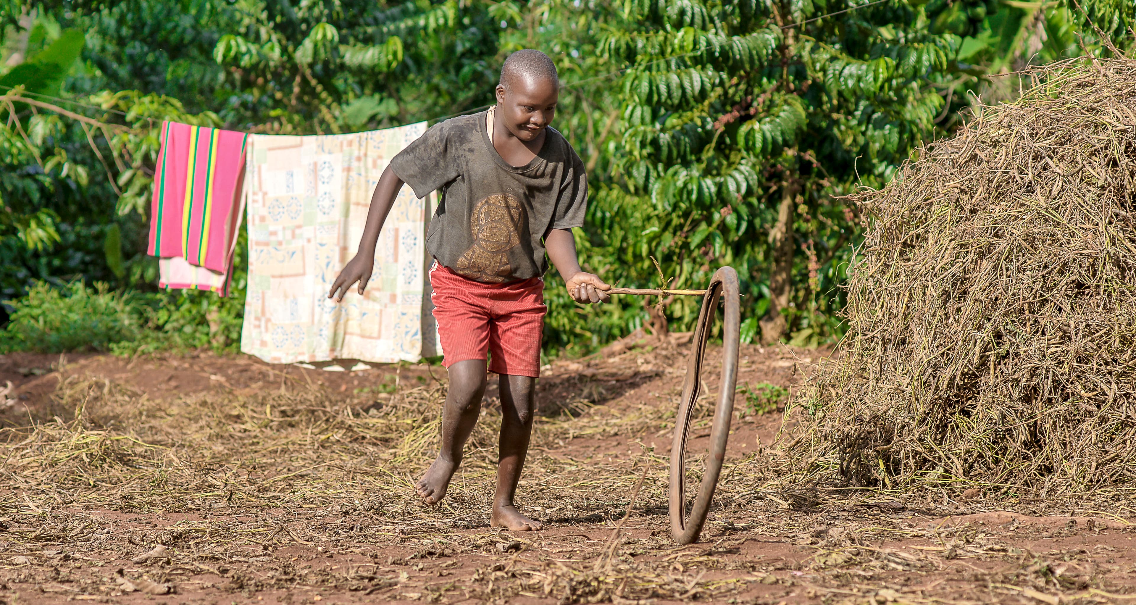 A shirtless boy runs outdoors past a clothesline of hanging laundry and a pile of dried grass.