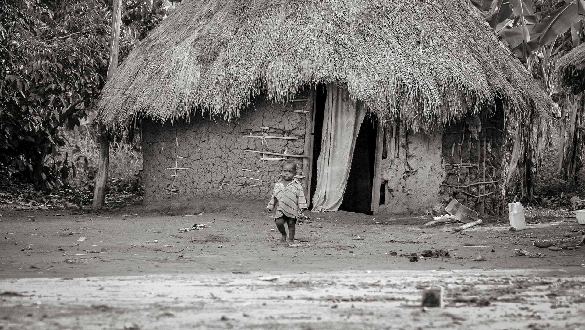 Black-and-white photo of a small thatched-roof mud hut with a child standing in the doorway.