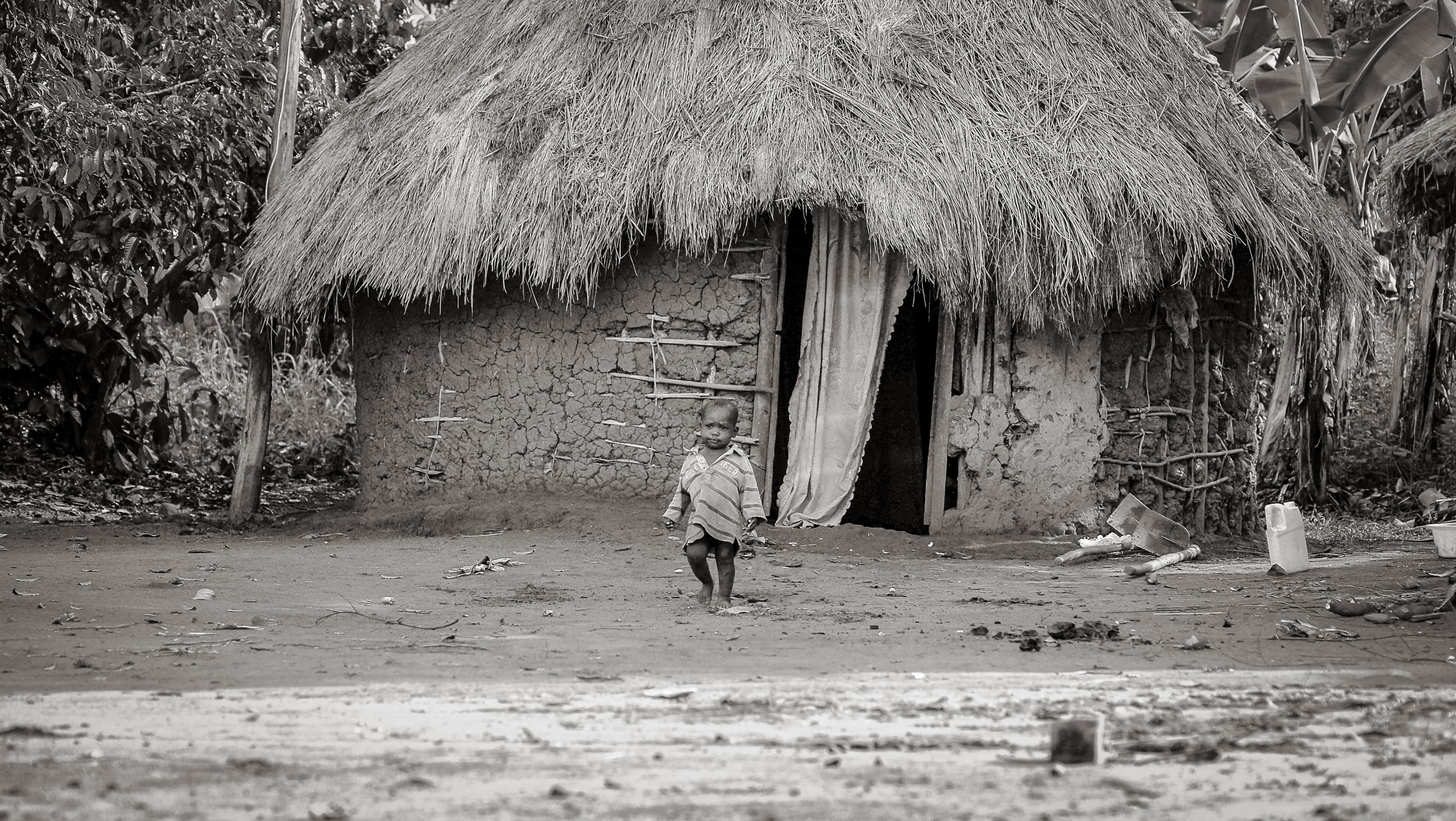Black-and-white photo of a small thatched-roof mud hut with a child standing in the doorway.