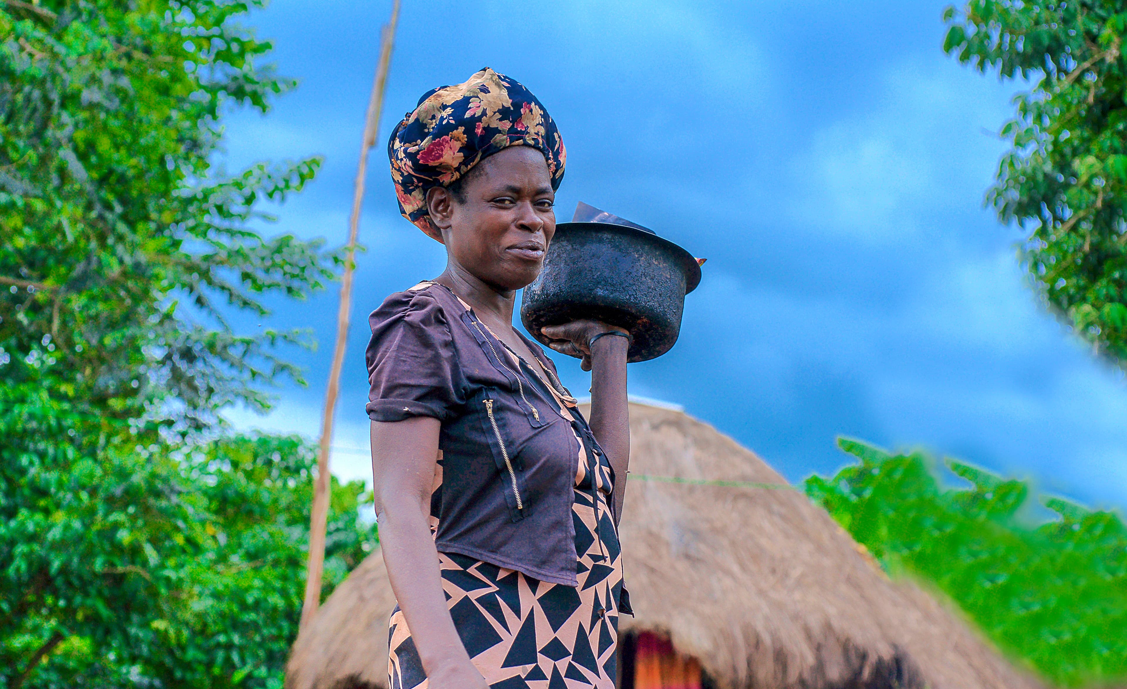 A woman in a patterned dress stands near a thatched hut holding a basin.