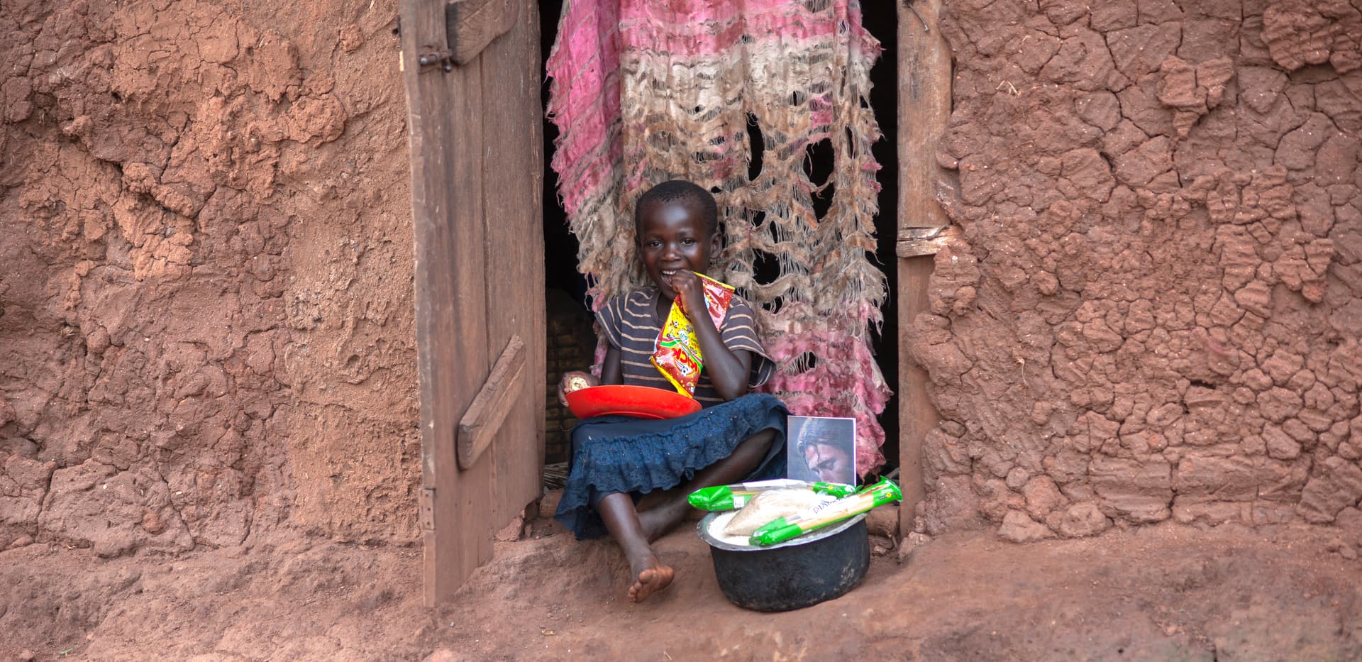 Young child sitting on the ground near a wooden doorway, holding a bowl with food items.