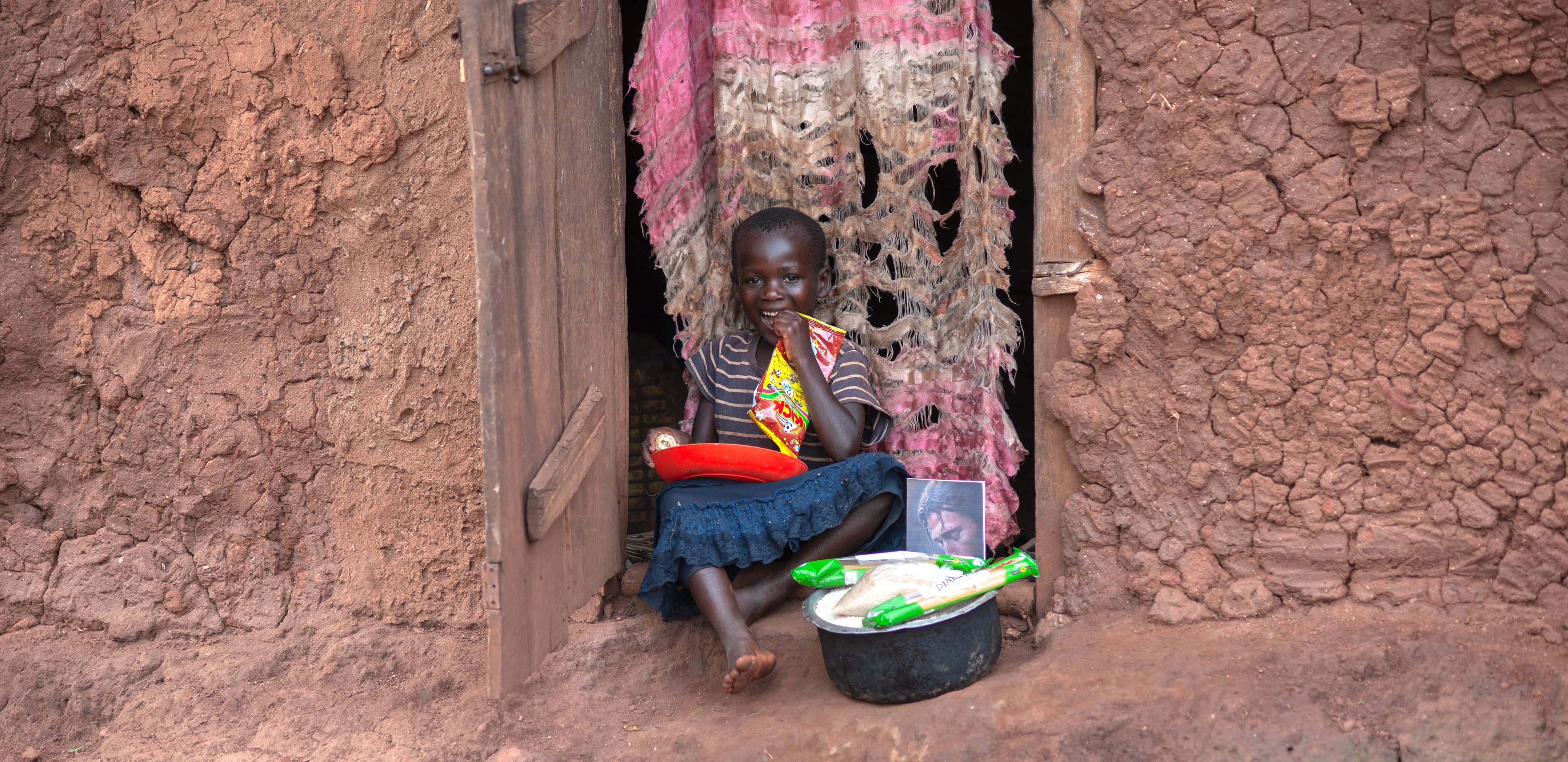 Young child sitting on the ground near a wooden doorway, holding a bowl with food items.