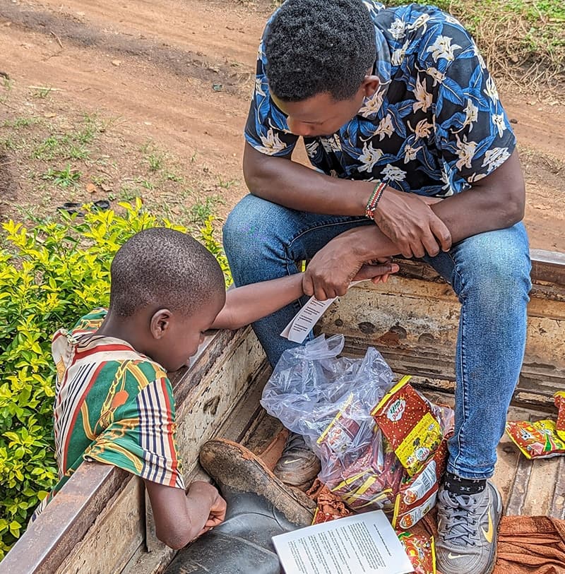 Man sitting outdoors reading with a boy, a bag of food gifts on the ground beside them.