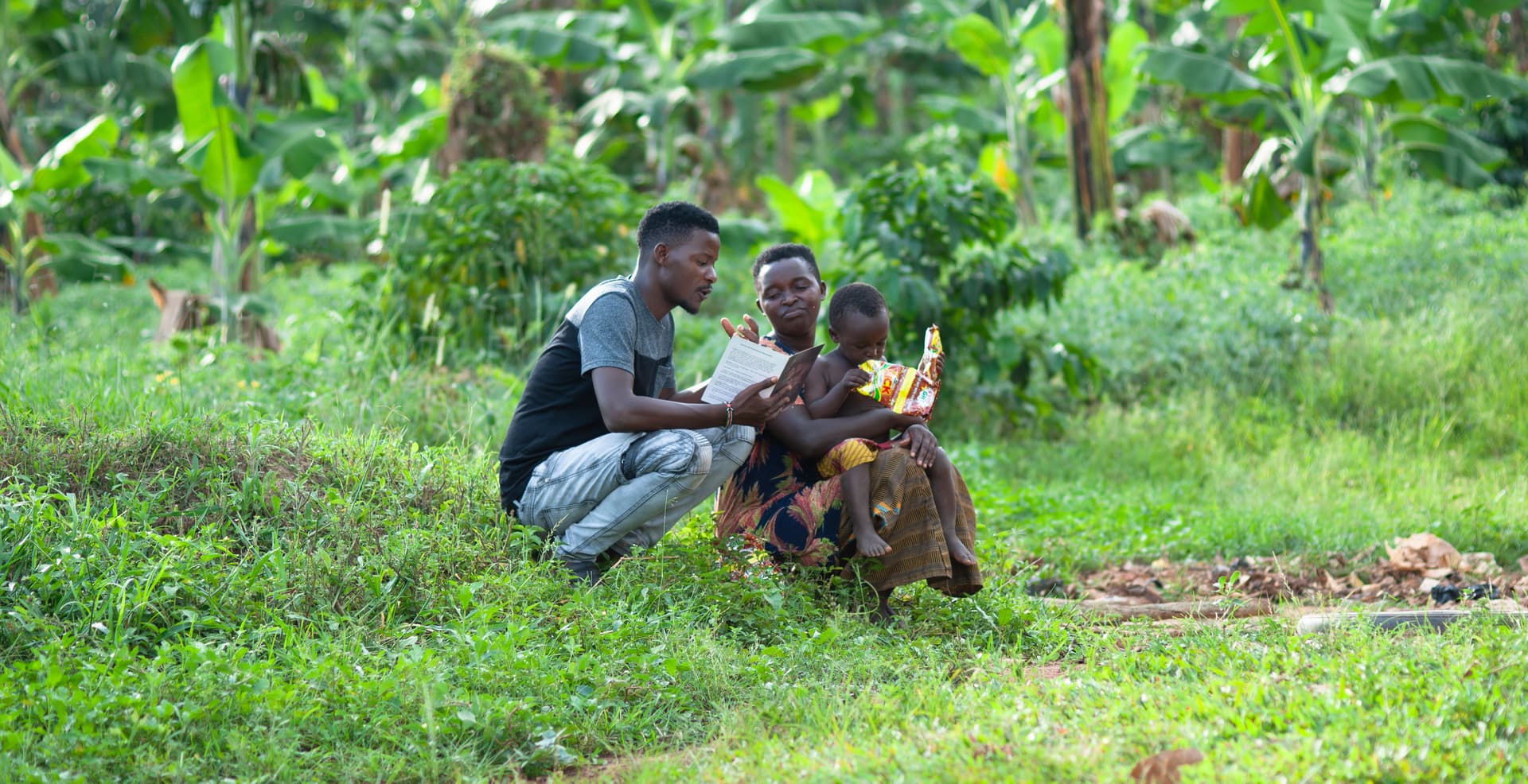 A man crouches in a grassy field talking with two young children.