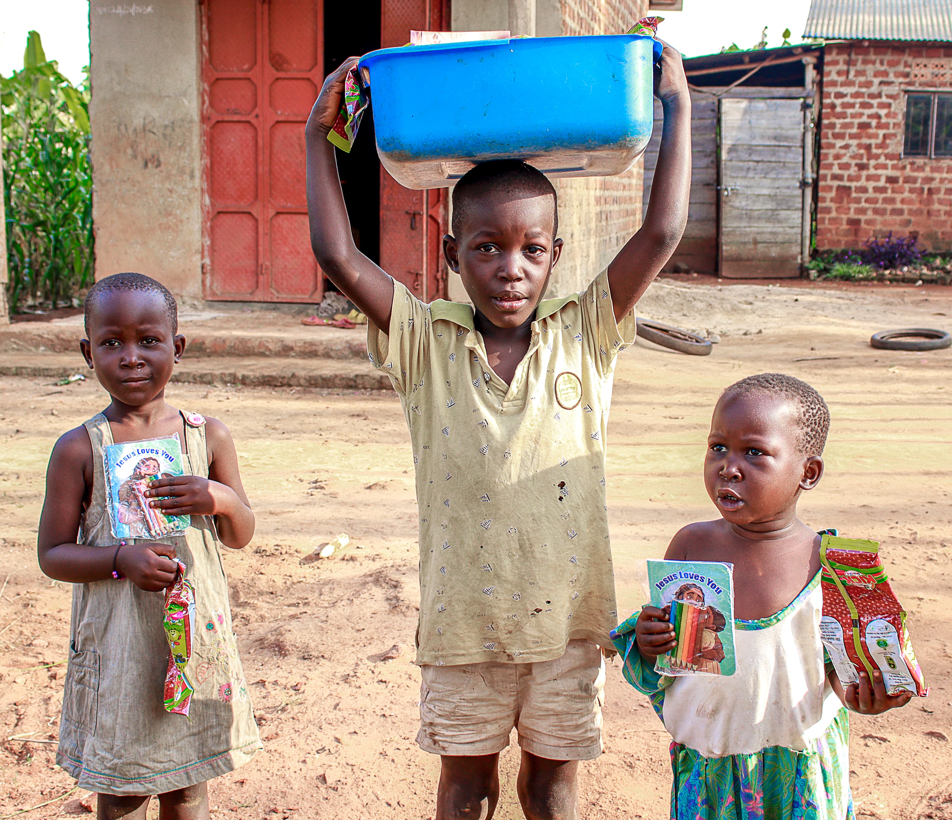 Three children stand outside a building with a red door; a boy holds a blue basin over his head while the others hold food packets and a gospel tract.