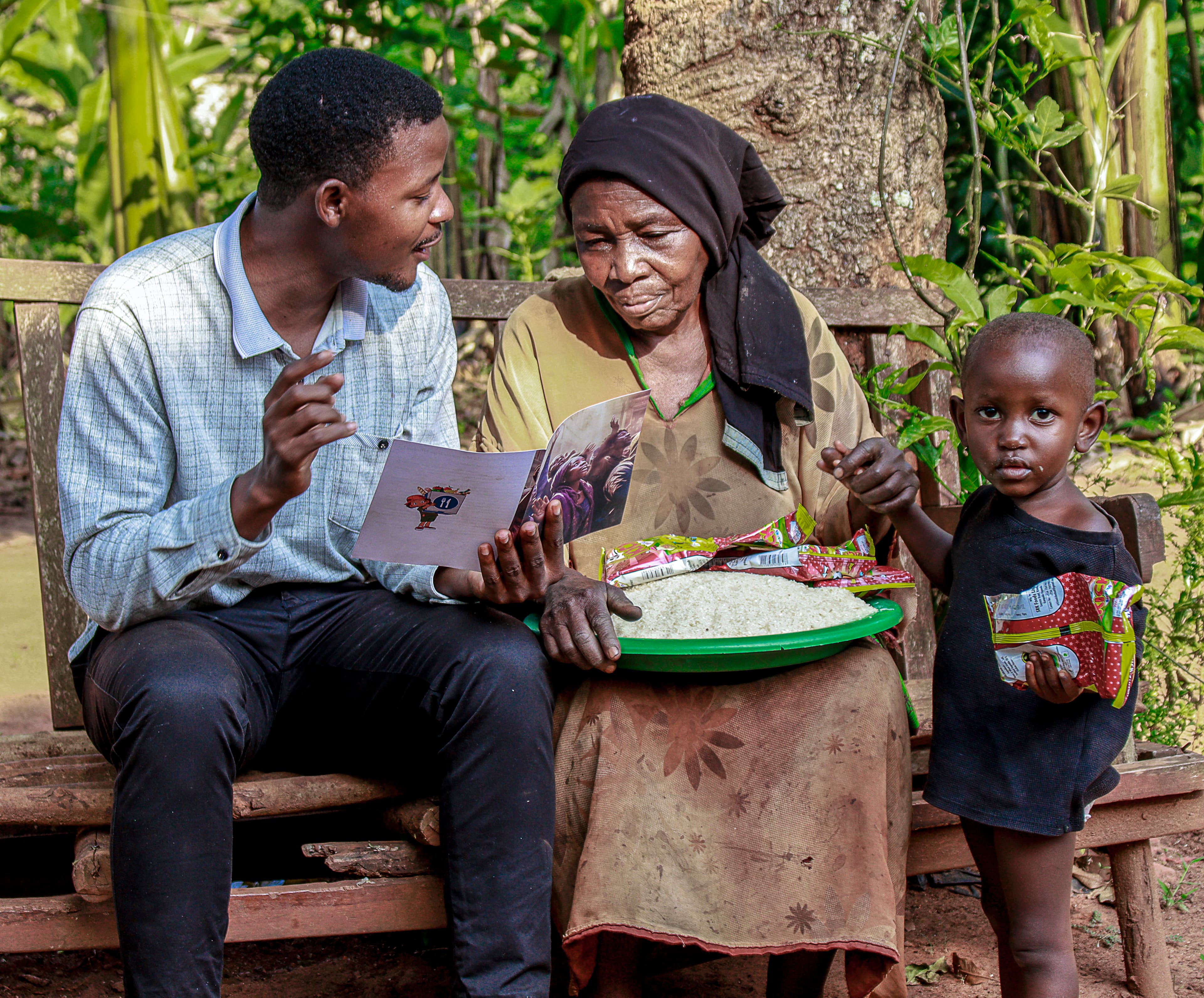 A man shares a gospel booklet with a woman and boy on a wooden bench, a green basin of food packets nearby.