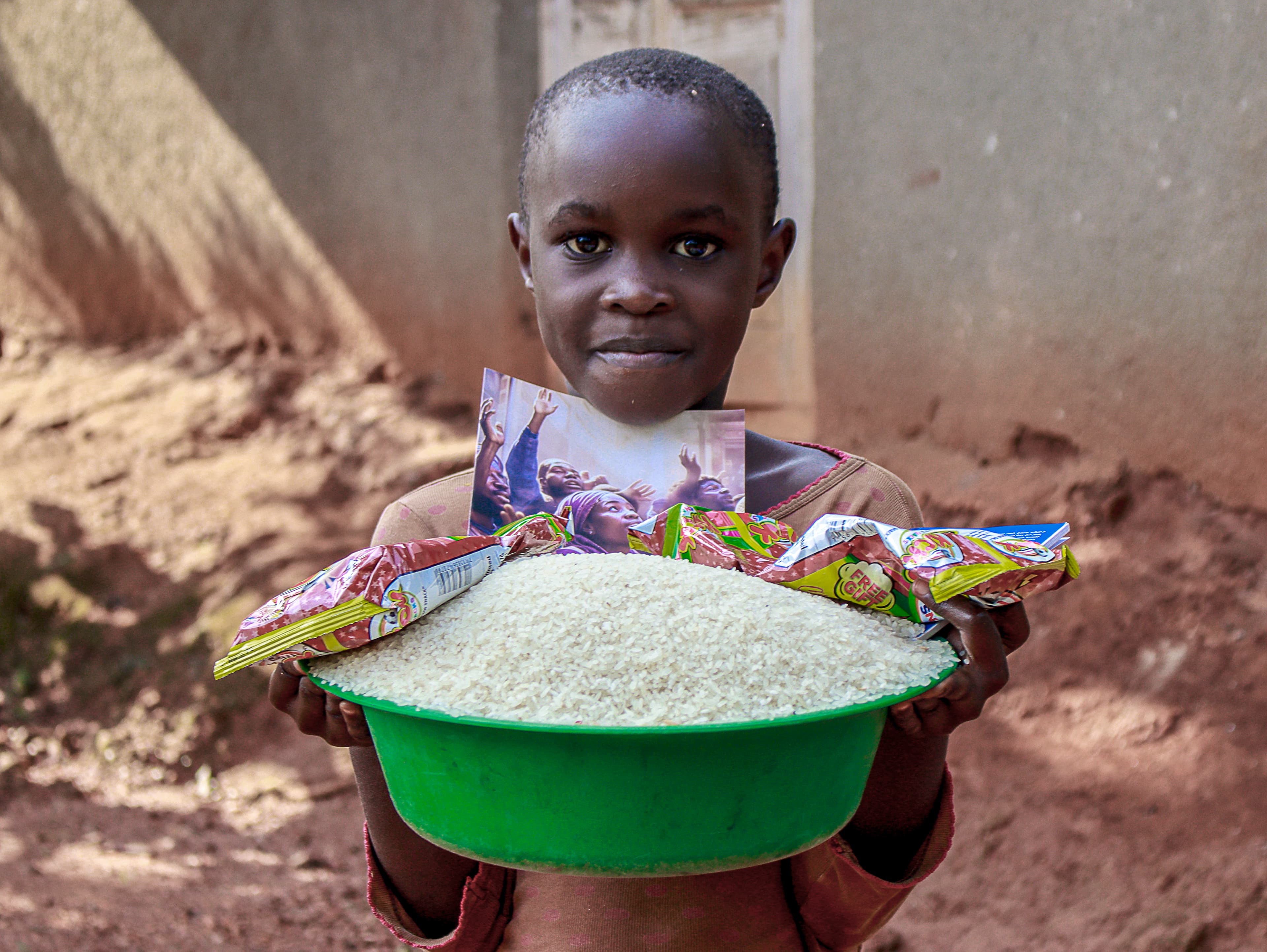 Close-up of a boy against a wall holding a green basin filled with rice