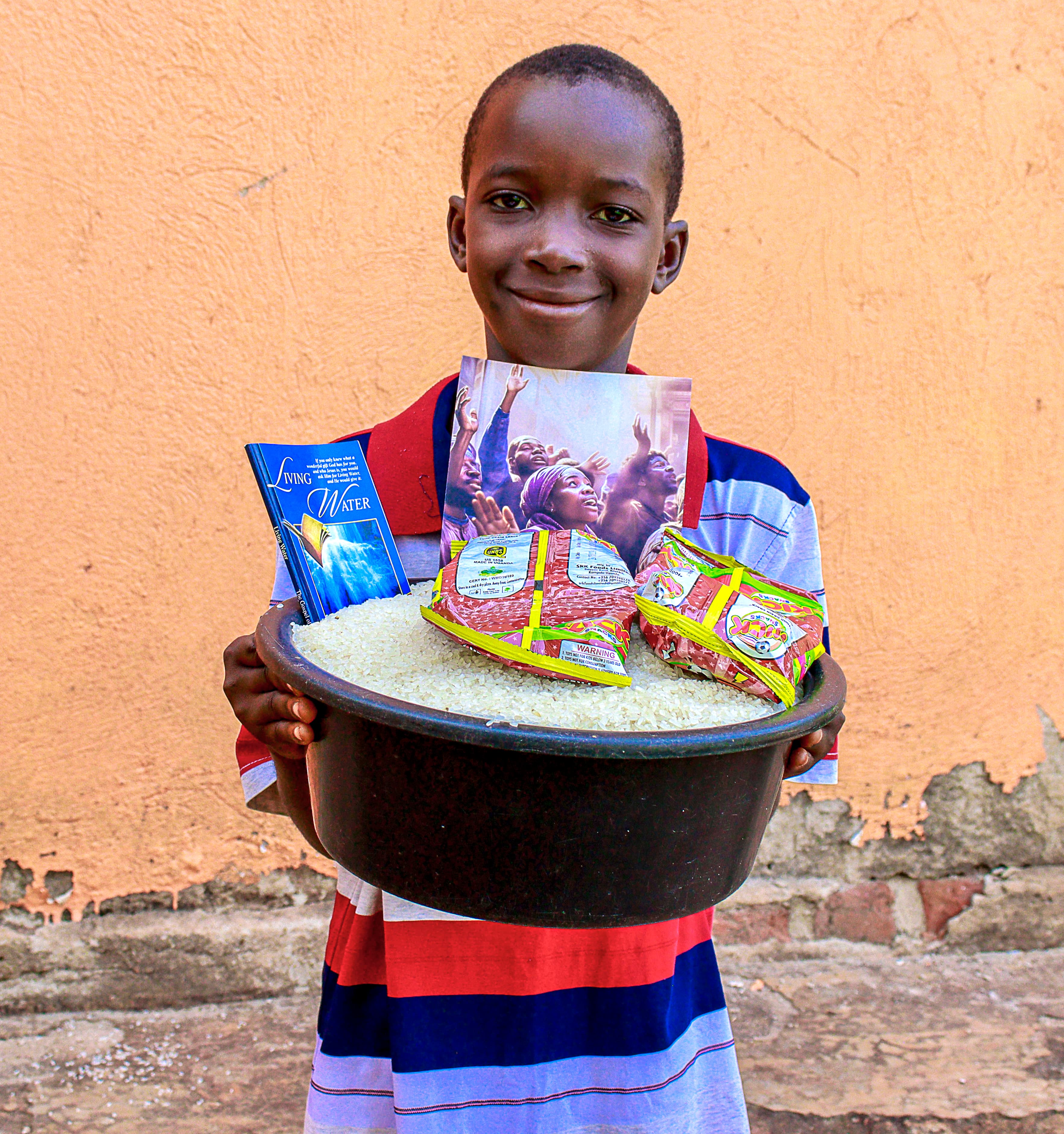 Boy smiling while holding a metal basin filled with food packets against an orange wall