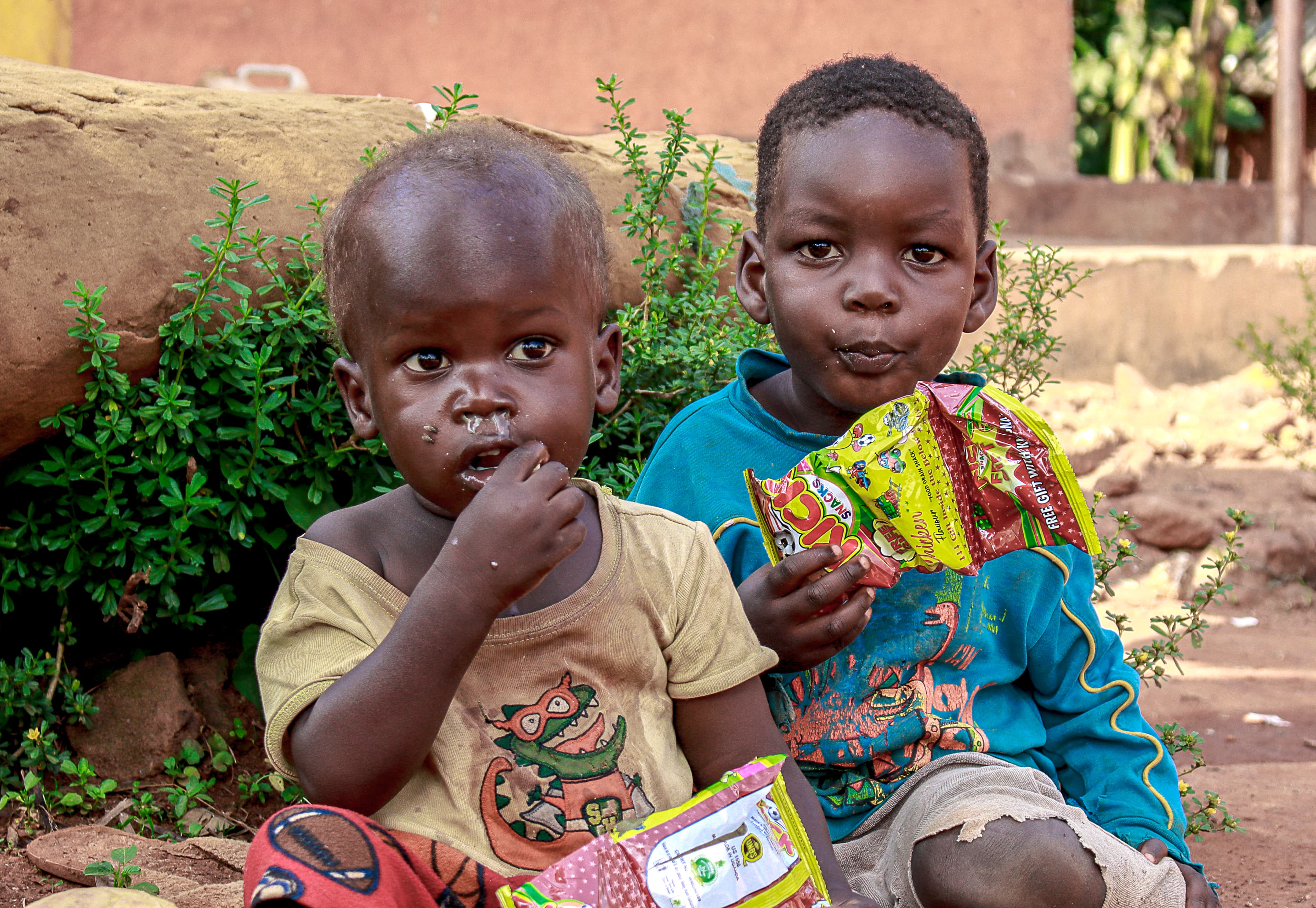 Two children sitting together outdoors, one eating a snack packet