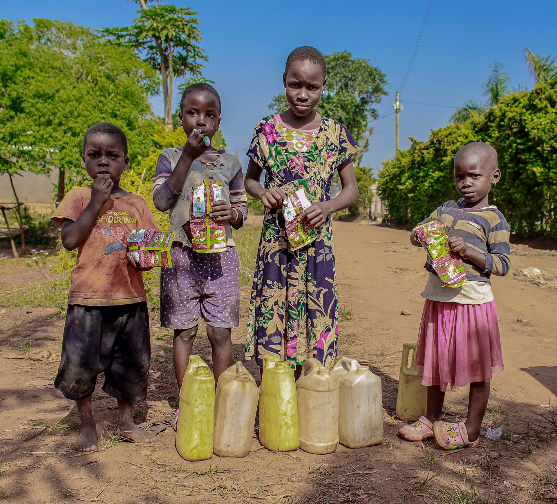 Four Ugandan children stand on a dirt road holding gospel tracts and food packets, with yellow water jerry cans lined up at their feet.