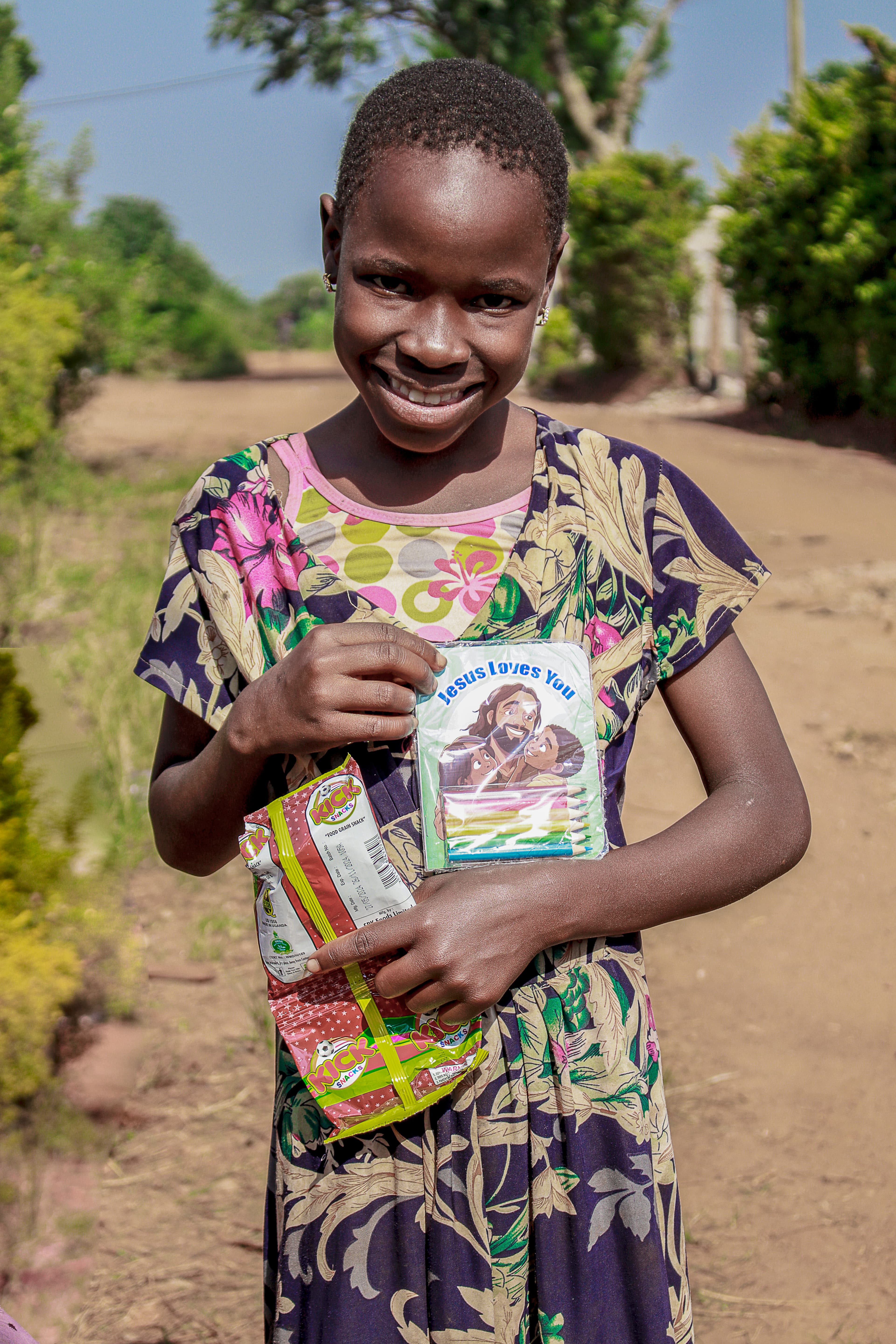 A girl in a dark floral dress smiles while holding a basket of food packets topped with a gospel tract.