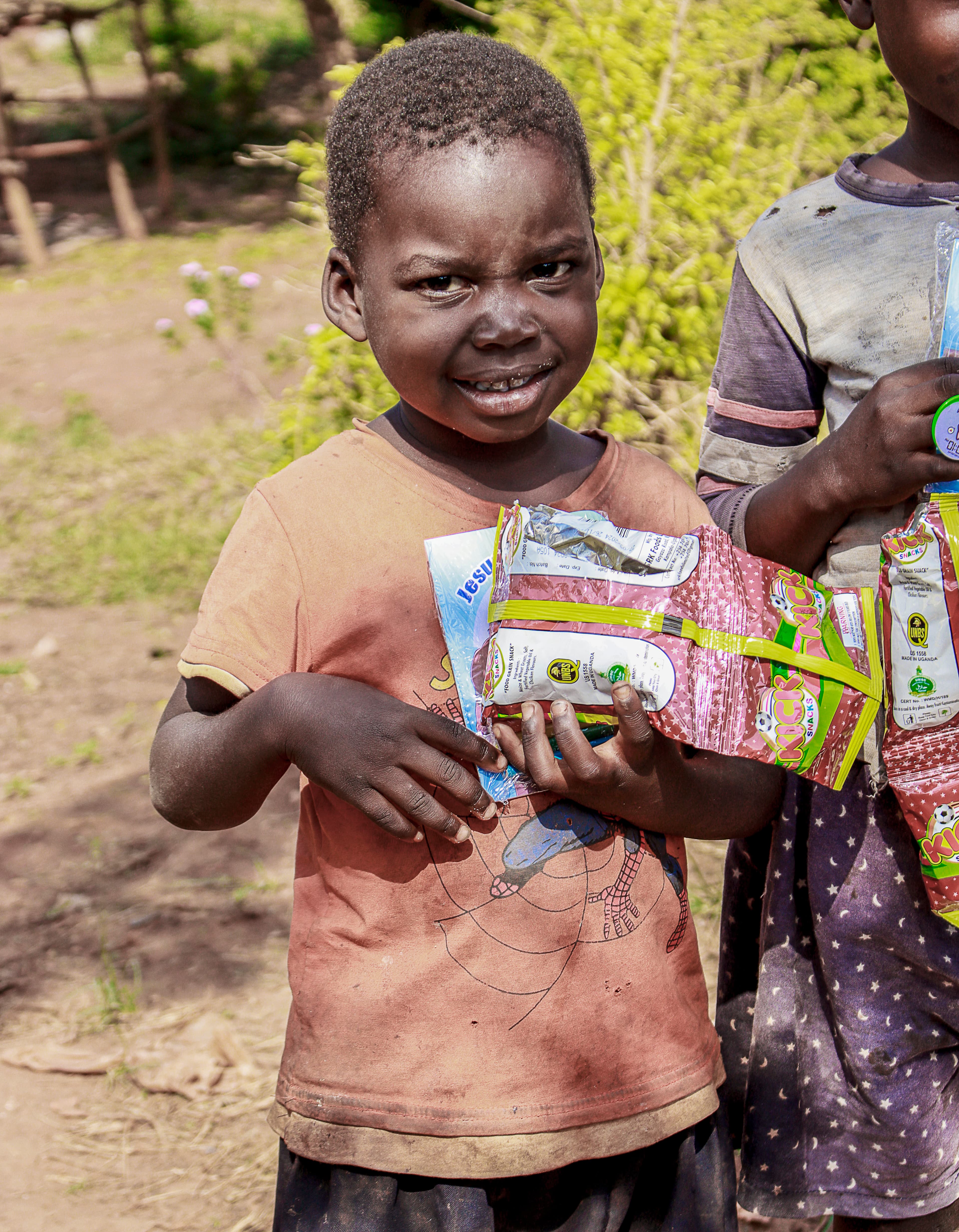 A boy in an orange shirt holds food packets and a gospel tract, with a girl in a blue dotted dress behind him.