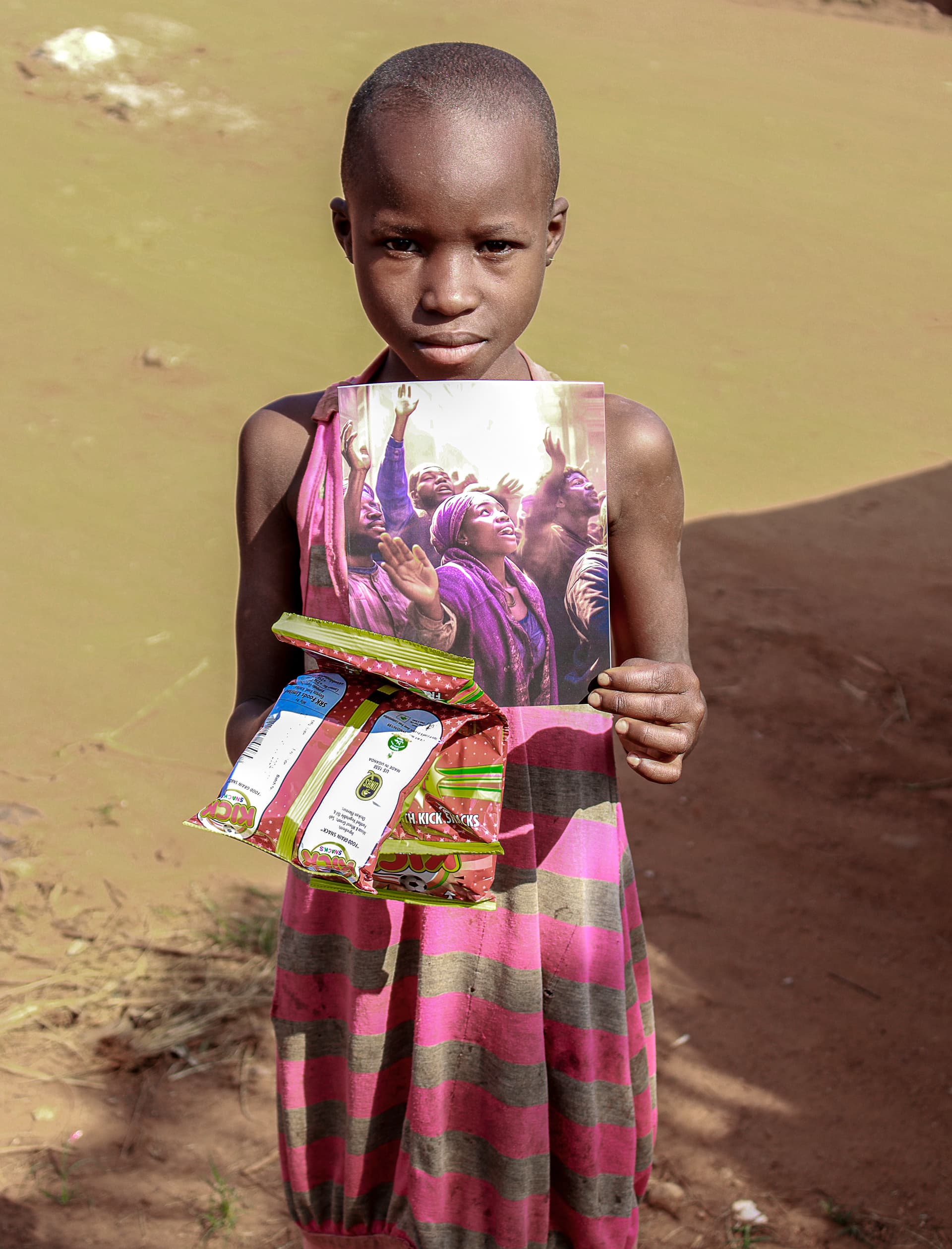 A girl in a magenta striped dress holds a food gift bundle wrapped with a gospel tract picture on a dirt path.