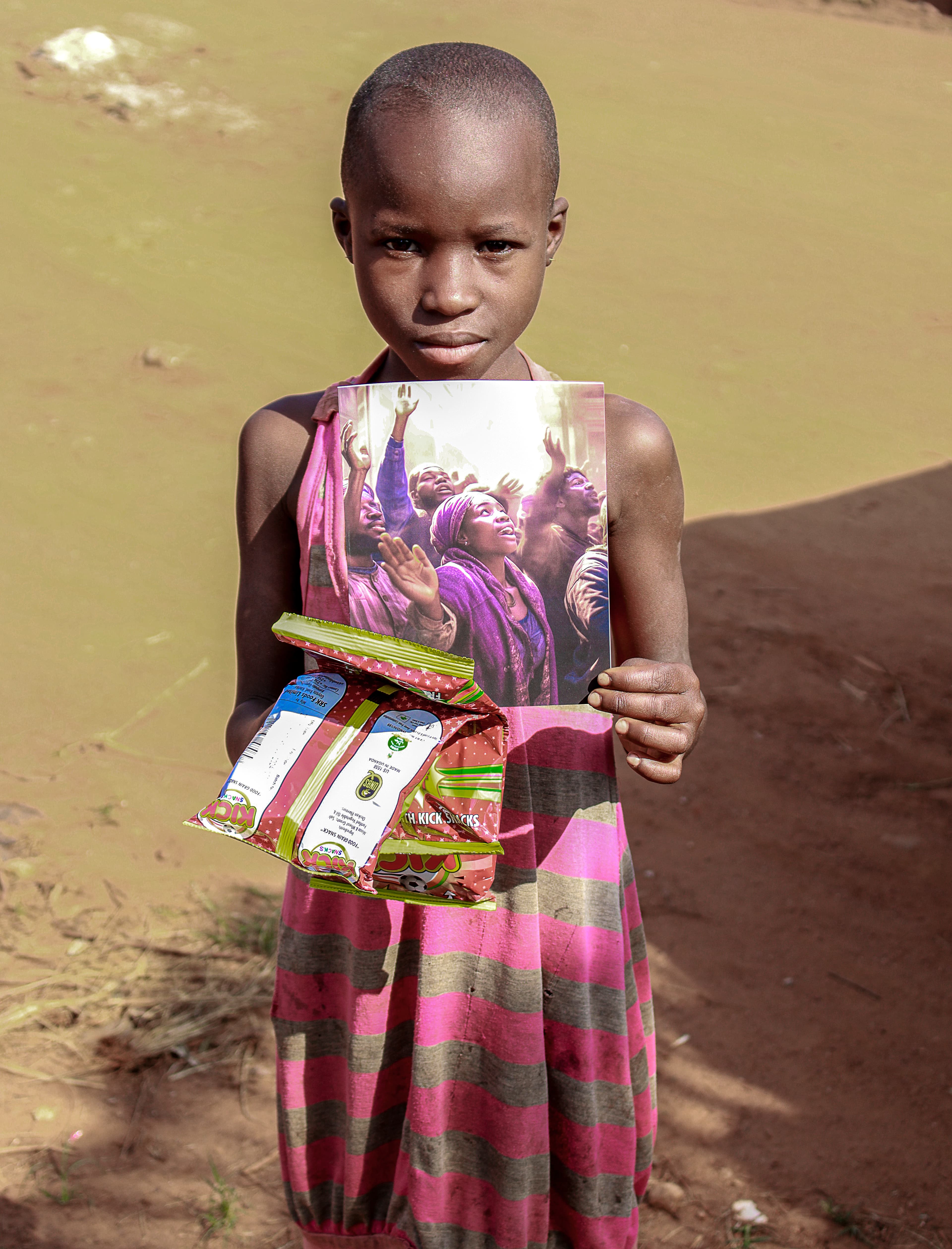 A girl in a magenta striped dress holds a food gift bundle wrapped with a gospel tract picture on a dirt path.