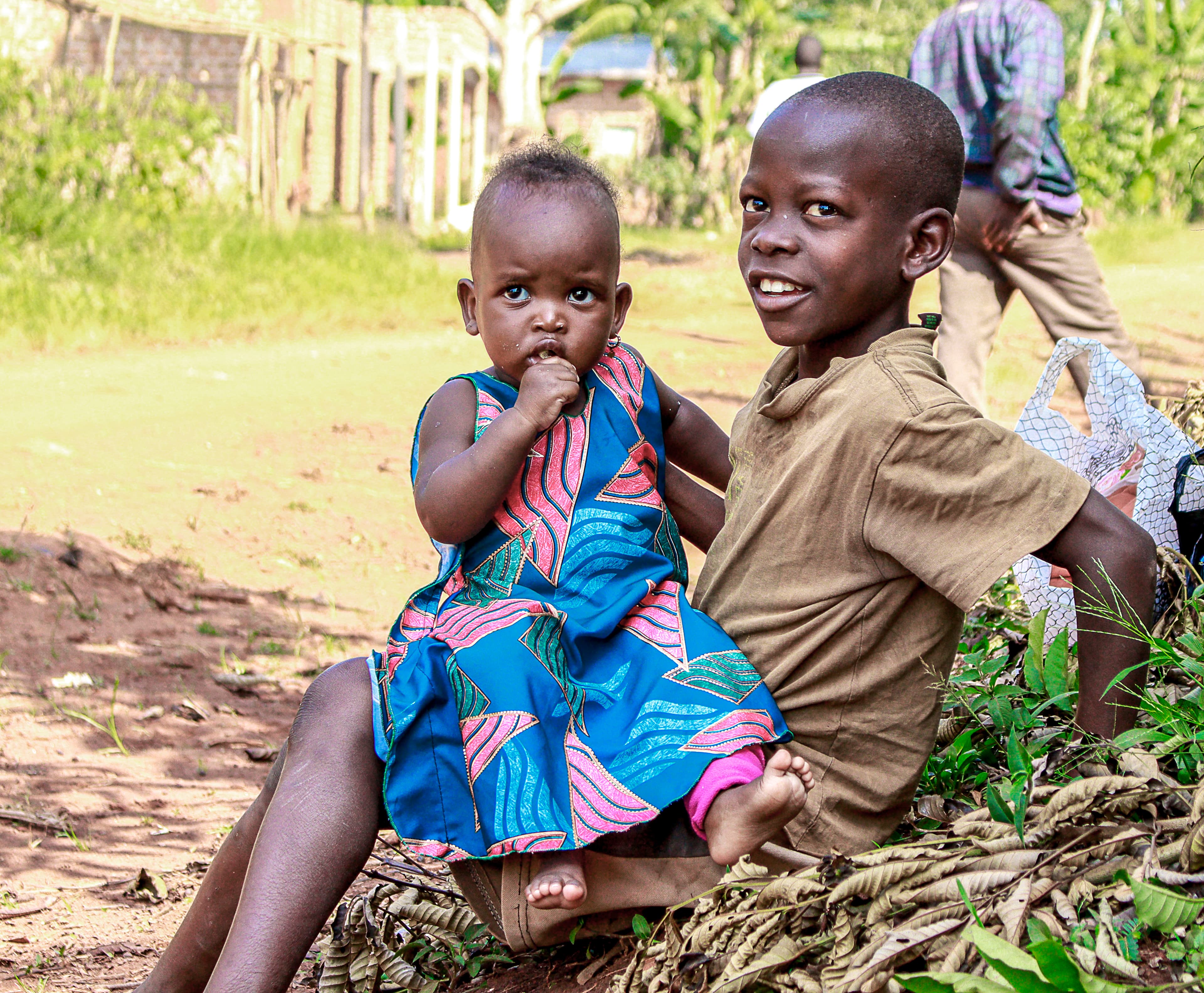 A toddler in a blue dress sits beside an older boy on a grassy hillside, with an adult's legs visible behind them.
