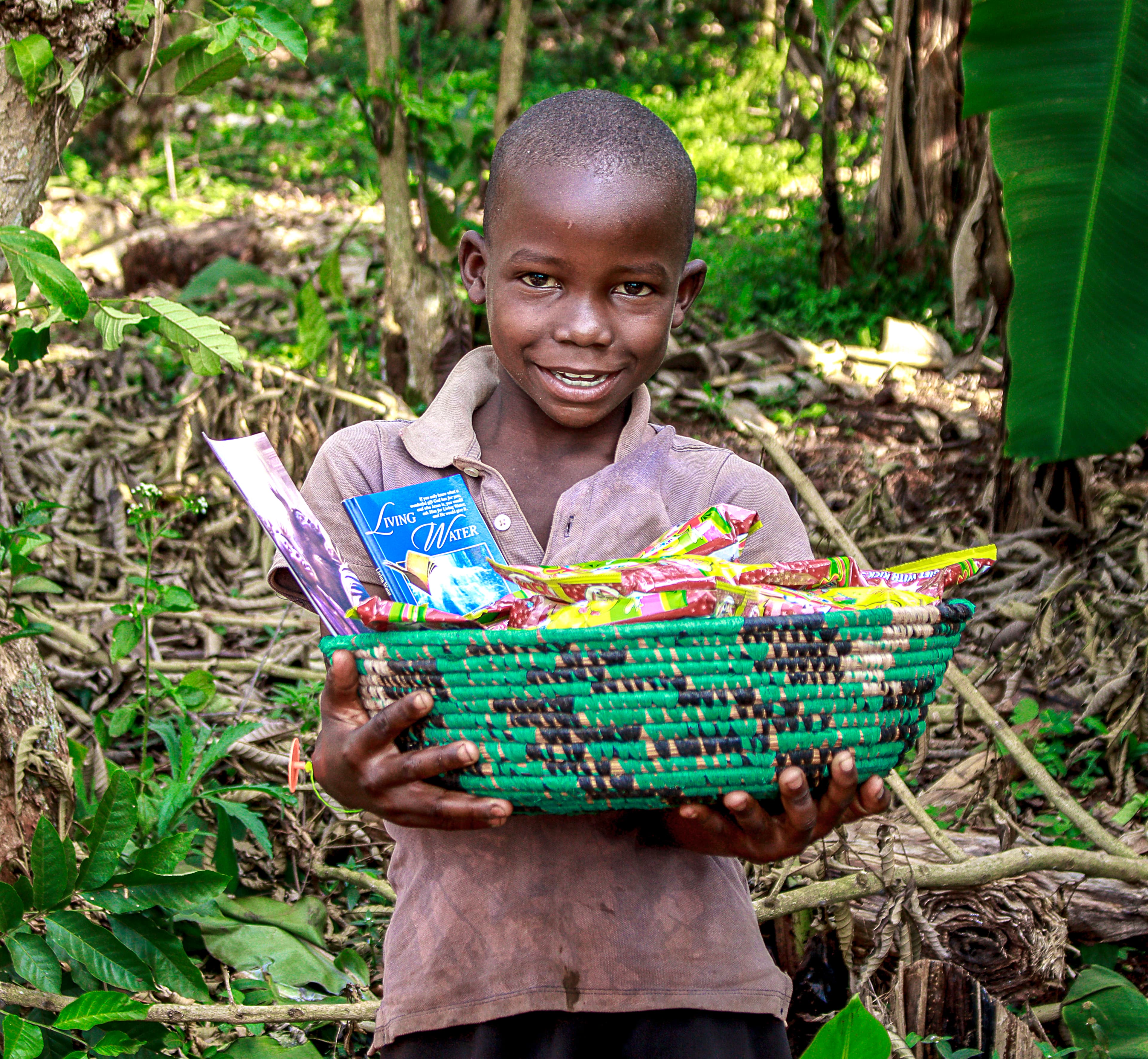 A boy stands among green foliage holding a woven basket of food packets topped with a gospel tract.