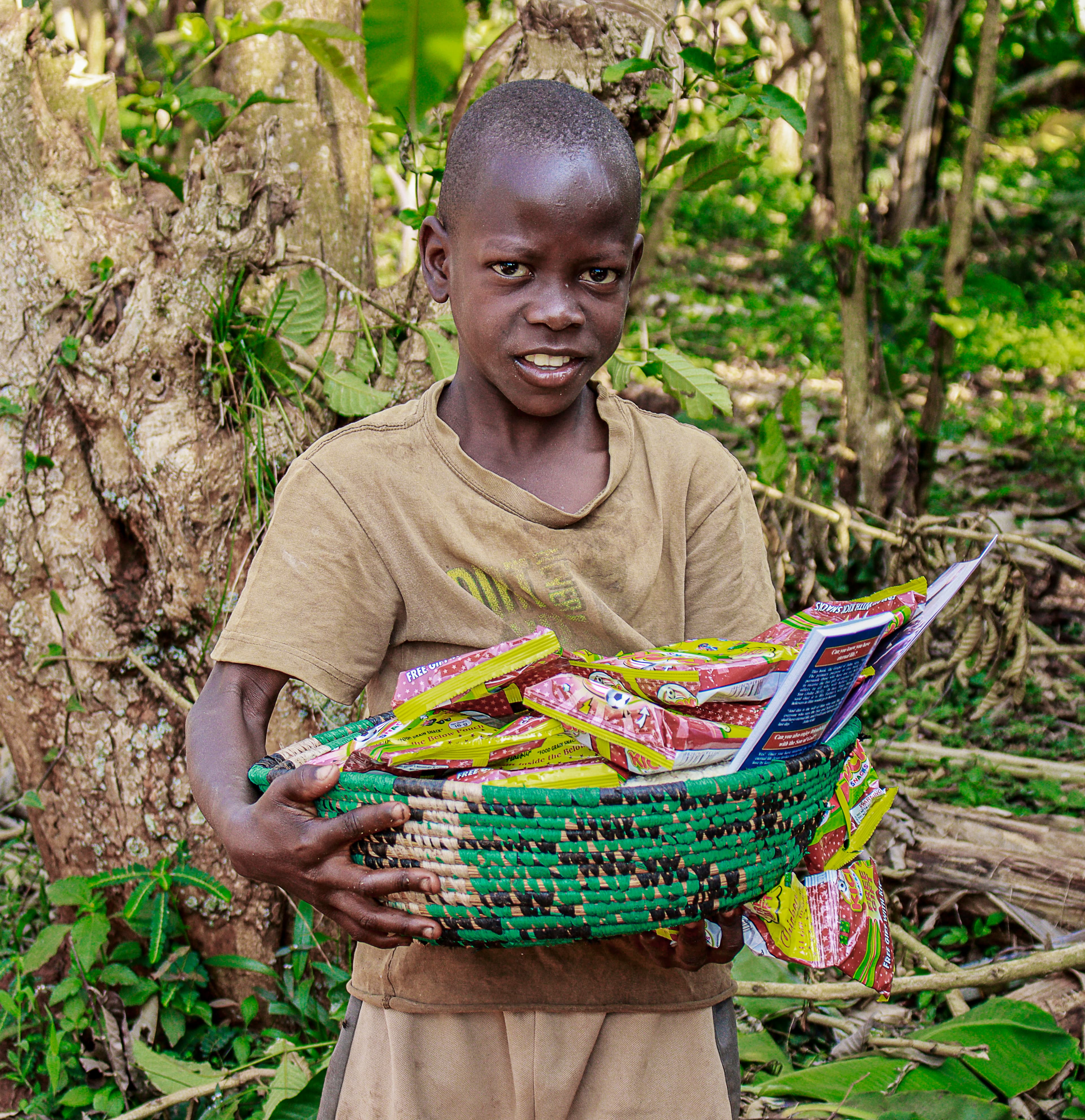 A boy in a tan shirt holds a basket of food packets and a gospel tract near a large tree.