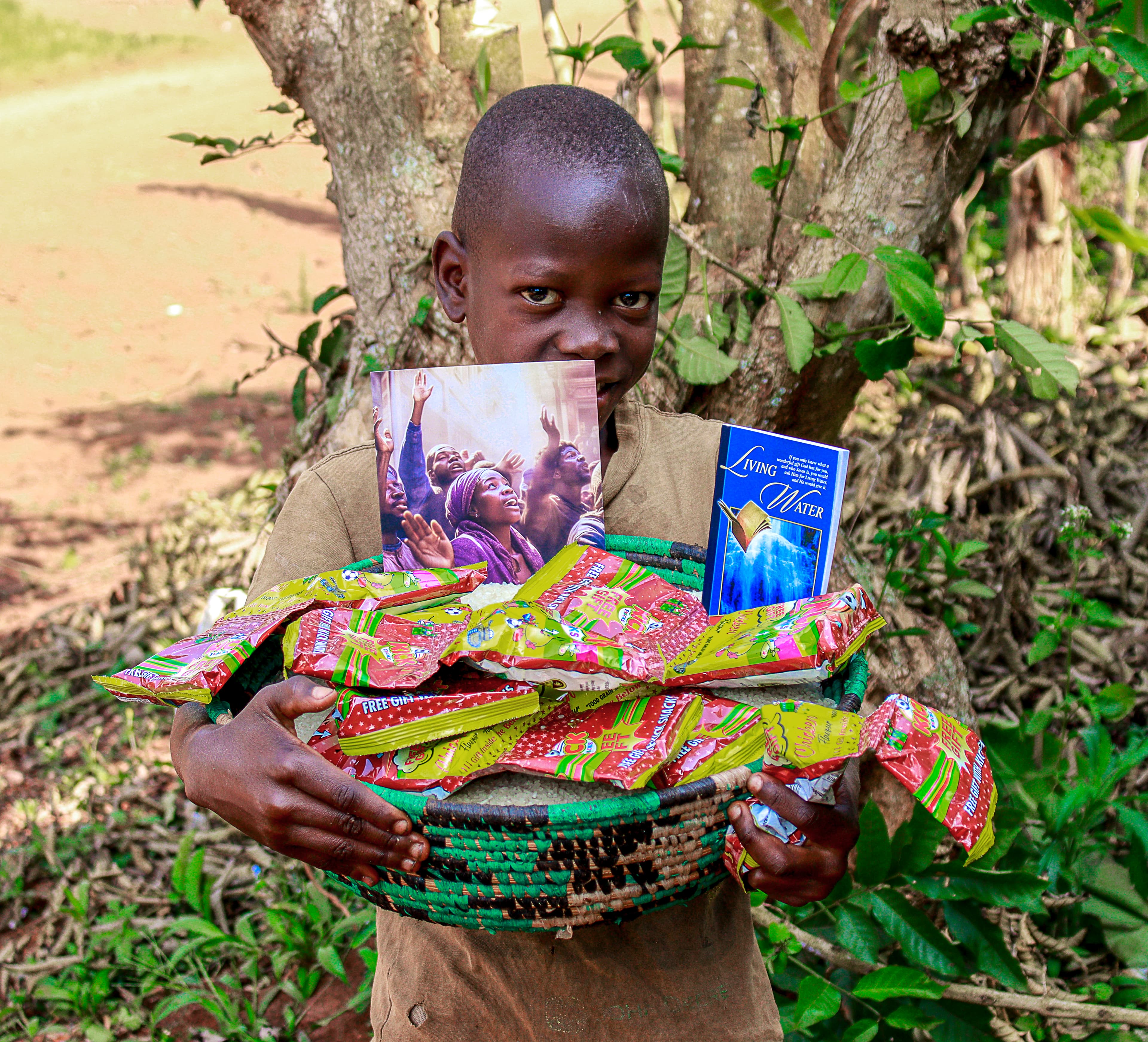 A boy holds a large bundle wrapped in colorful fabric that partly covers his face, standing among trees.