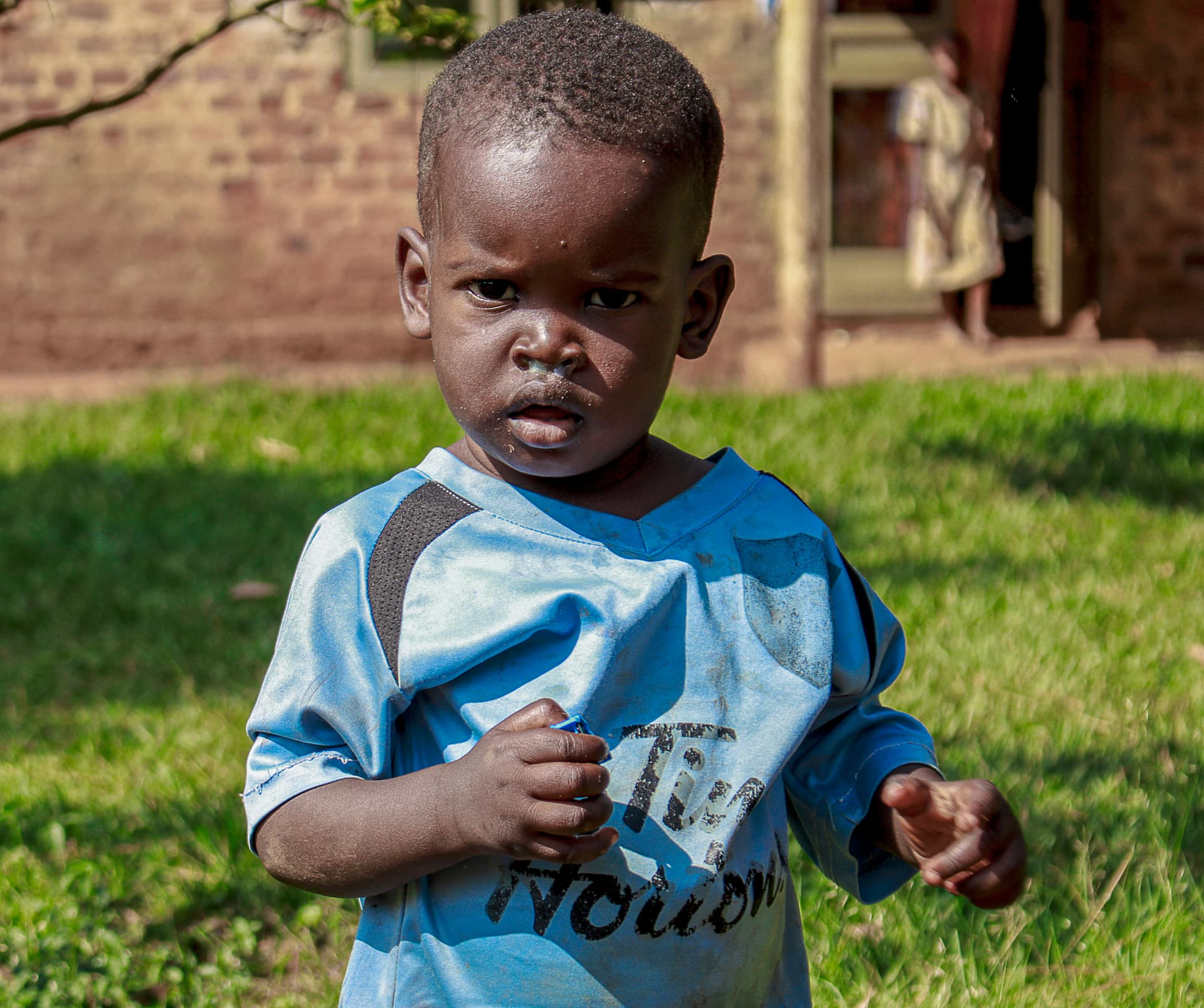 A young boy in a blue shirt stands alone on grass in front of a house.