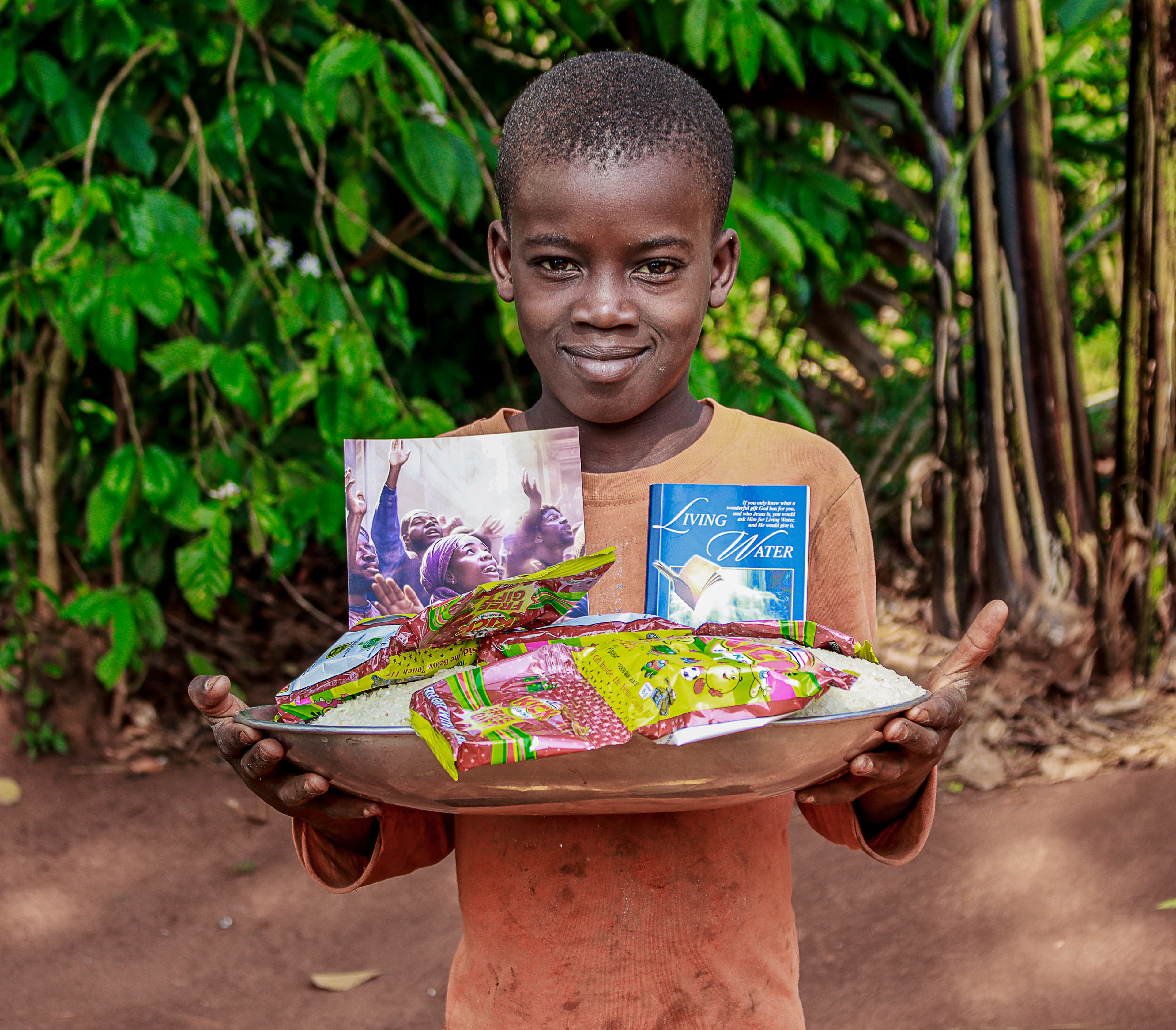 A boy in a rust-orange shirt smiles while holding a woven basket of food packets and a gospel tract.