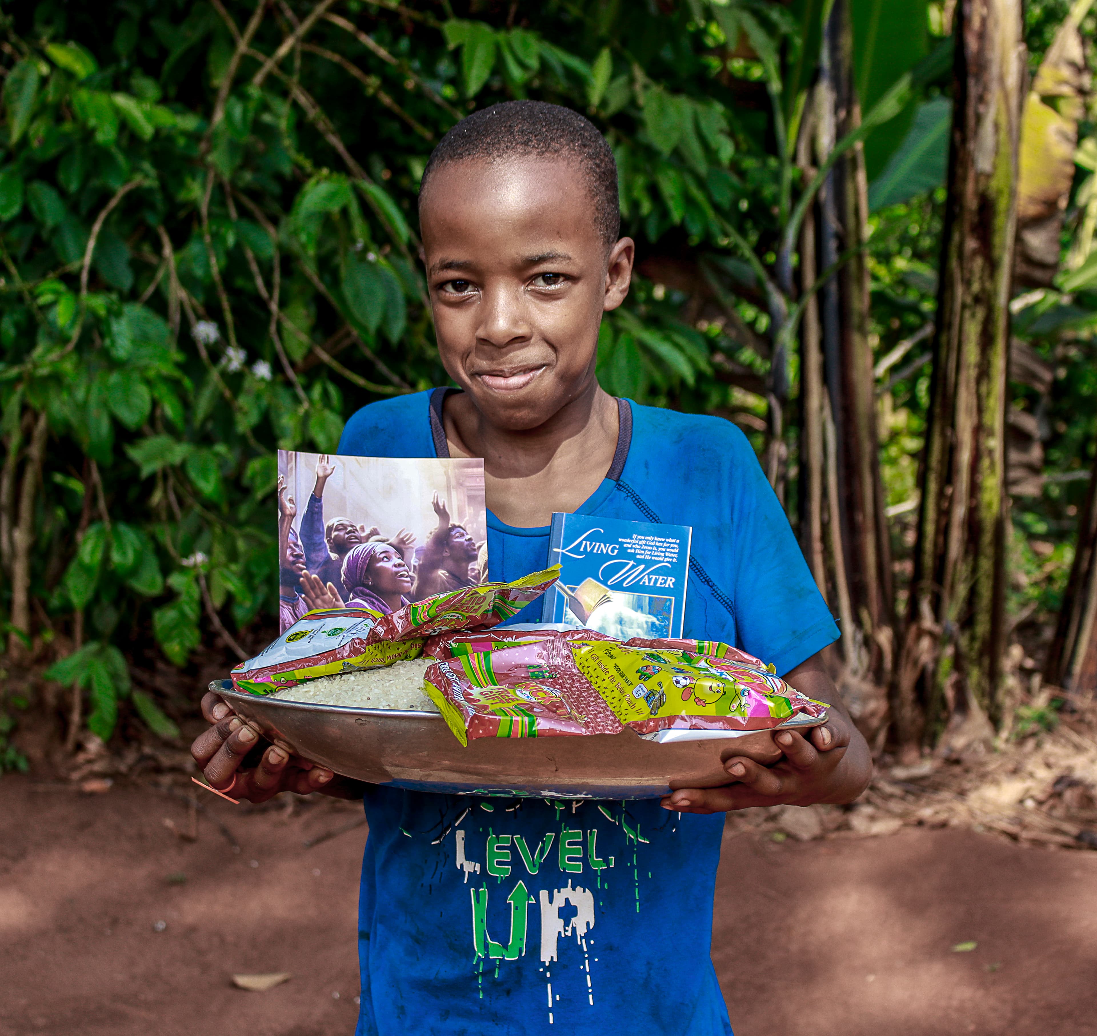 A boy in a blue "Level Up" T-shirt holds a basin with a gospel tract and food packets.