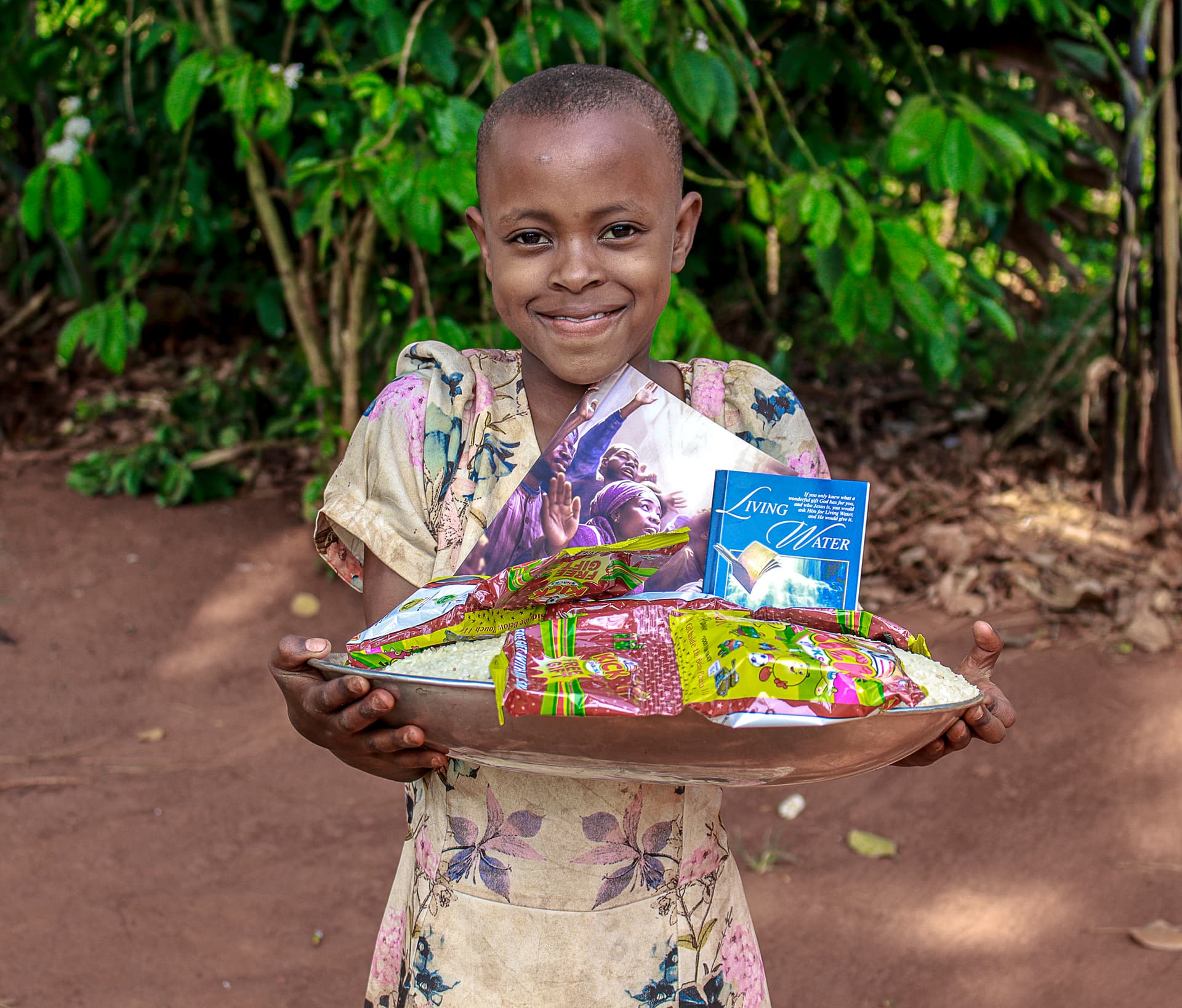 A smiling boy holds a white basin containing a gospel tract and food packets in a wooded clearing.