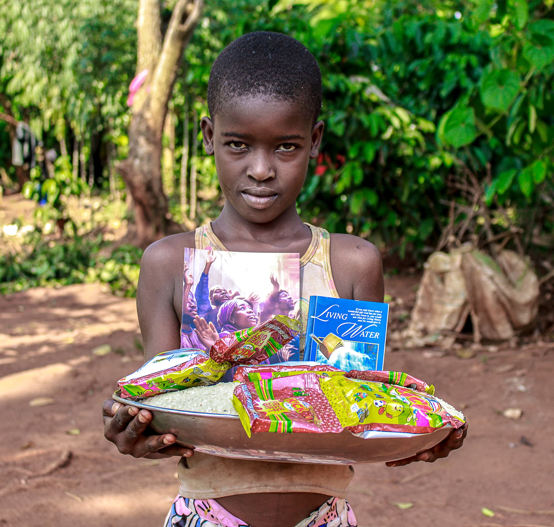 A girl holds a plate of food packets and a gospel tract while standing in a garden.