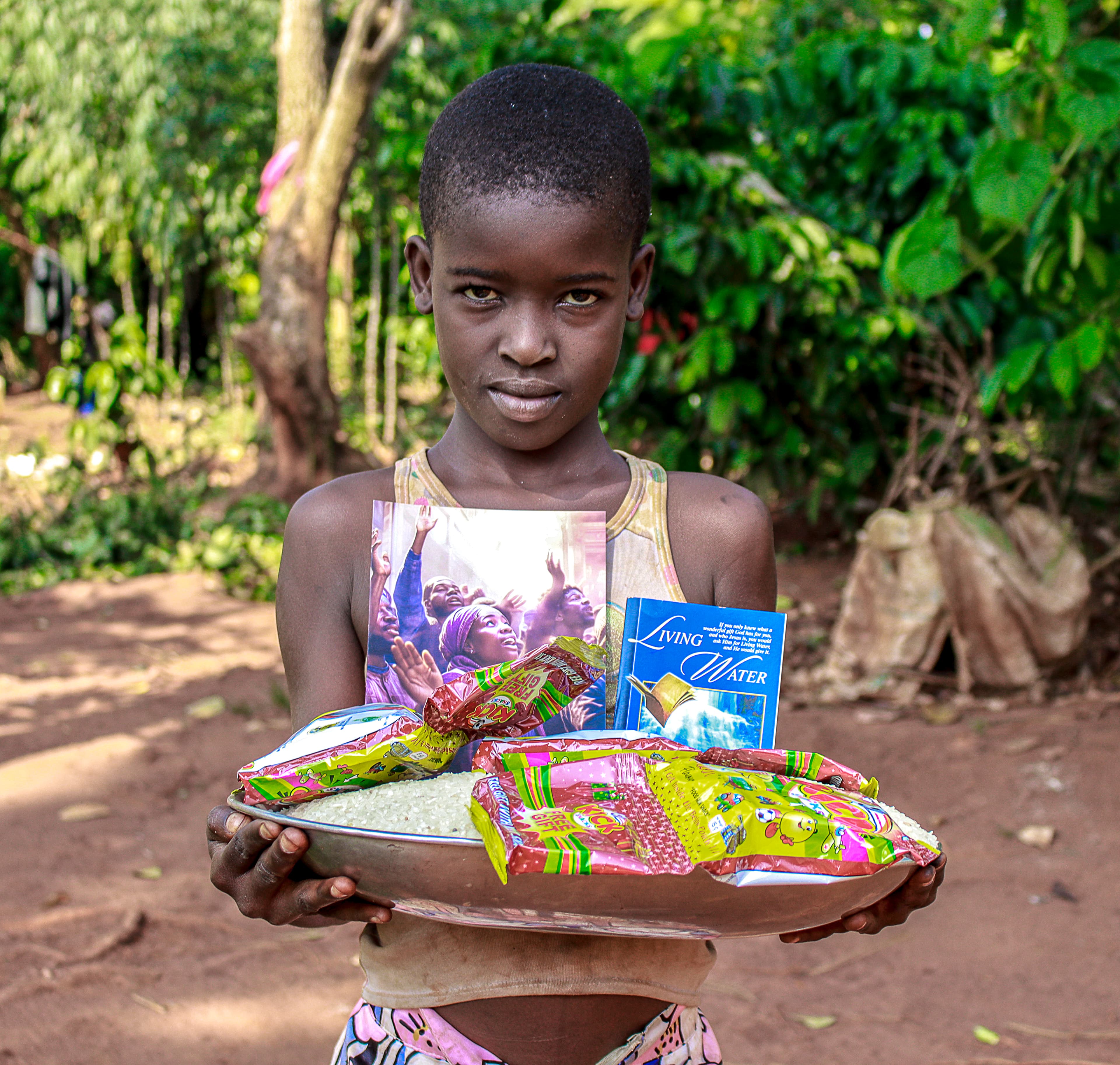 A girl holds a plate of food packets and a gospel tract while standing in a garden.