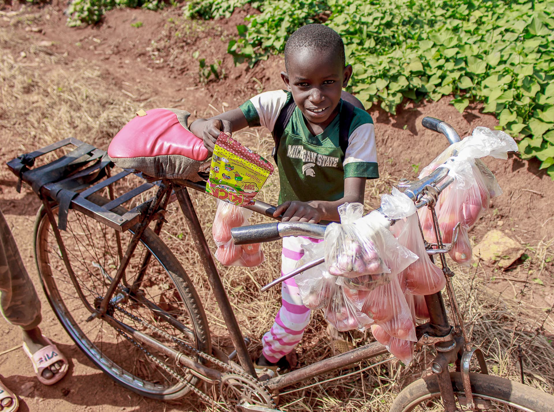 A boy stands beside a bicycle loaded with pink plastic bags of supplies on a dirt road.