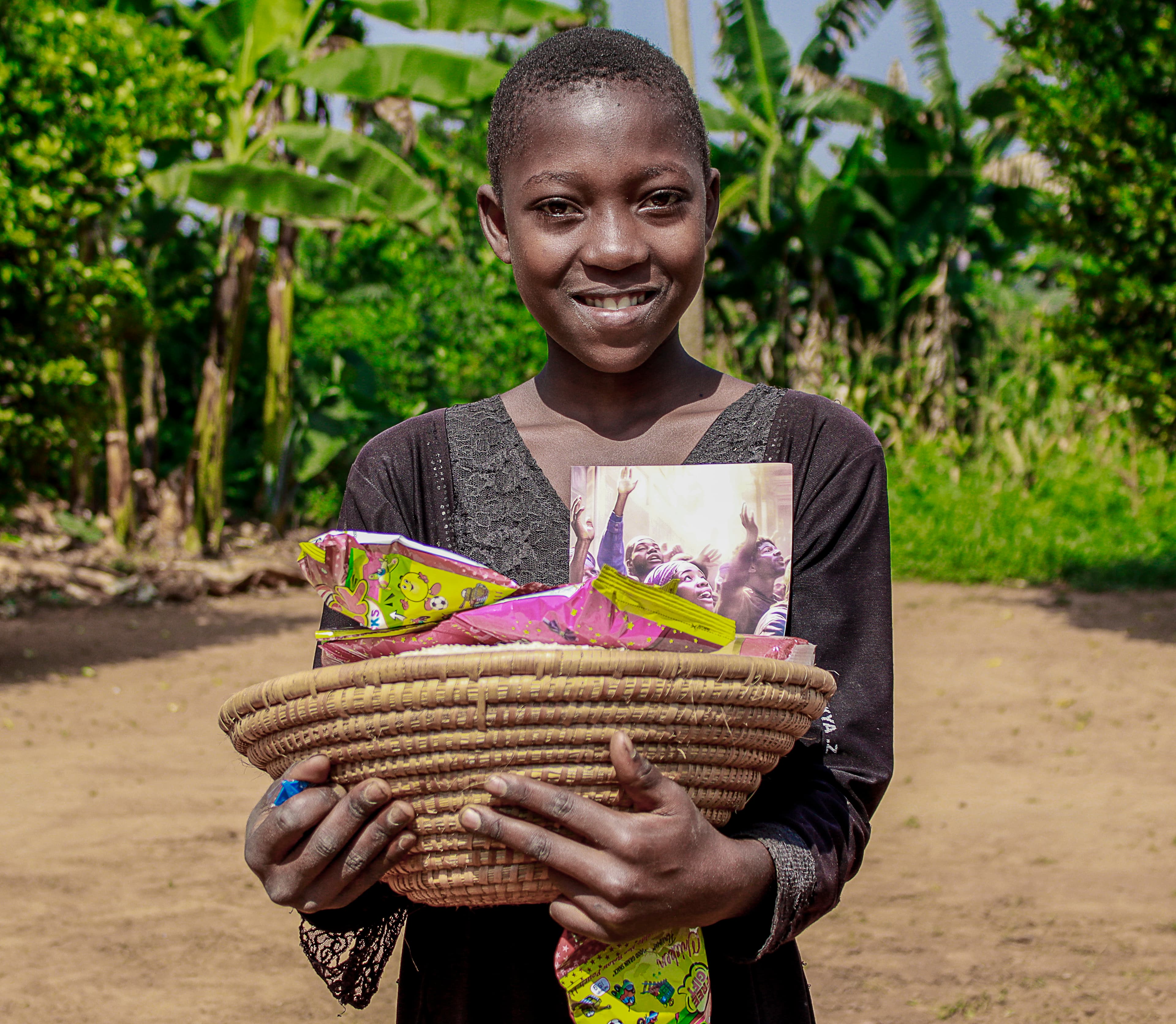 A boy smiles while holding a woven basket of food packets and a gospel tract on a dirt path.