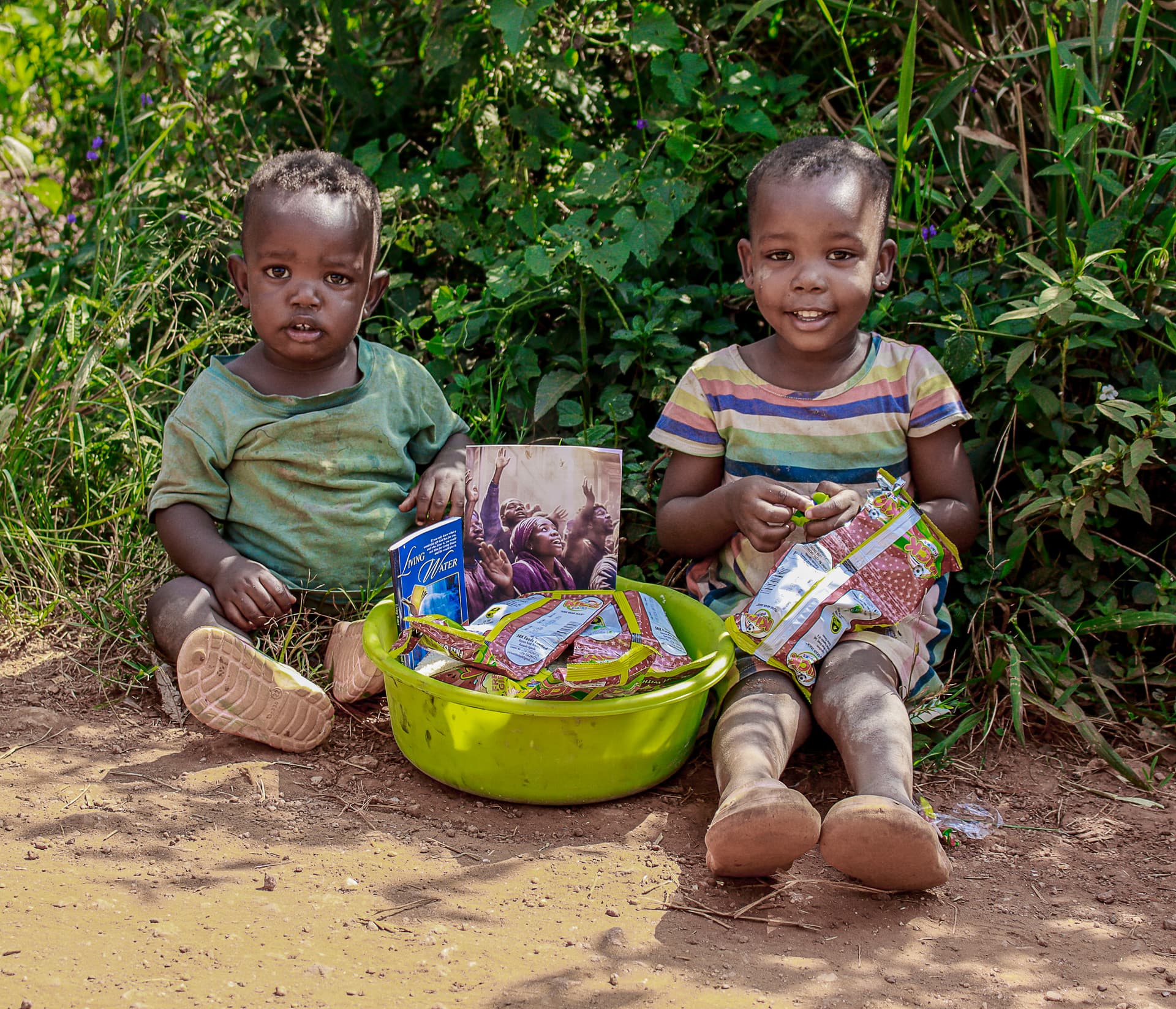Two young children sit on the ground beside a green basin filled with food packets and gospel tracts.