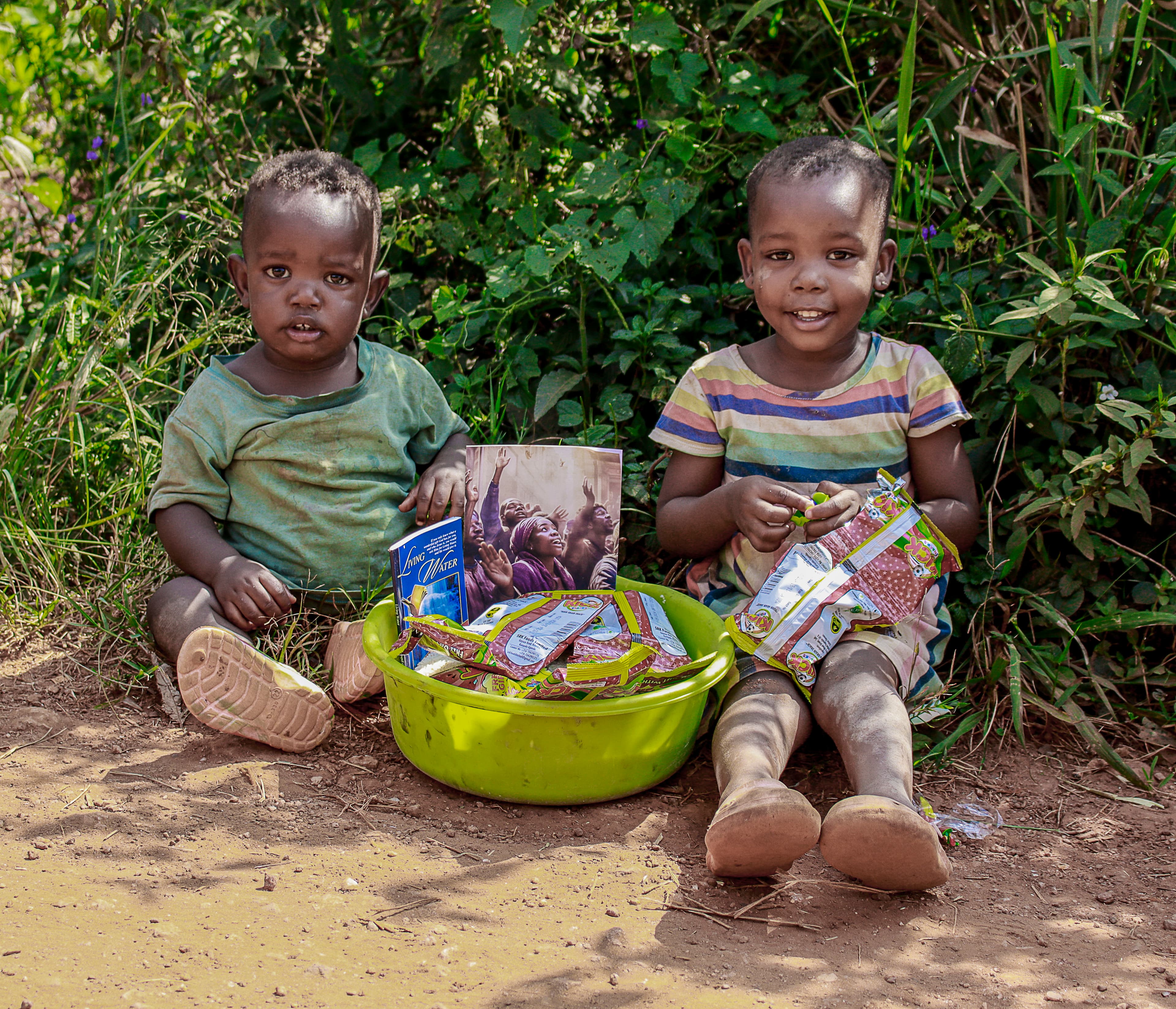 Two young children sit on the ground beside a green basin filled with food packets and gospel tracts.