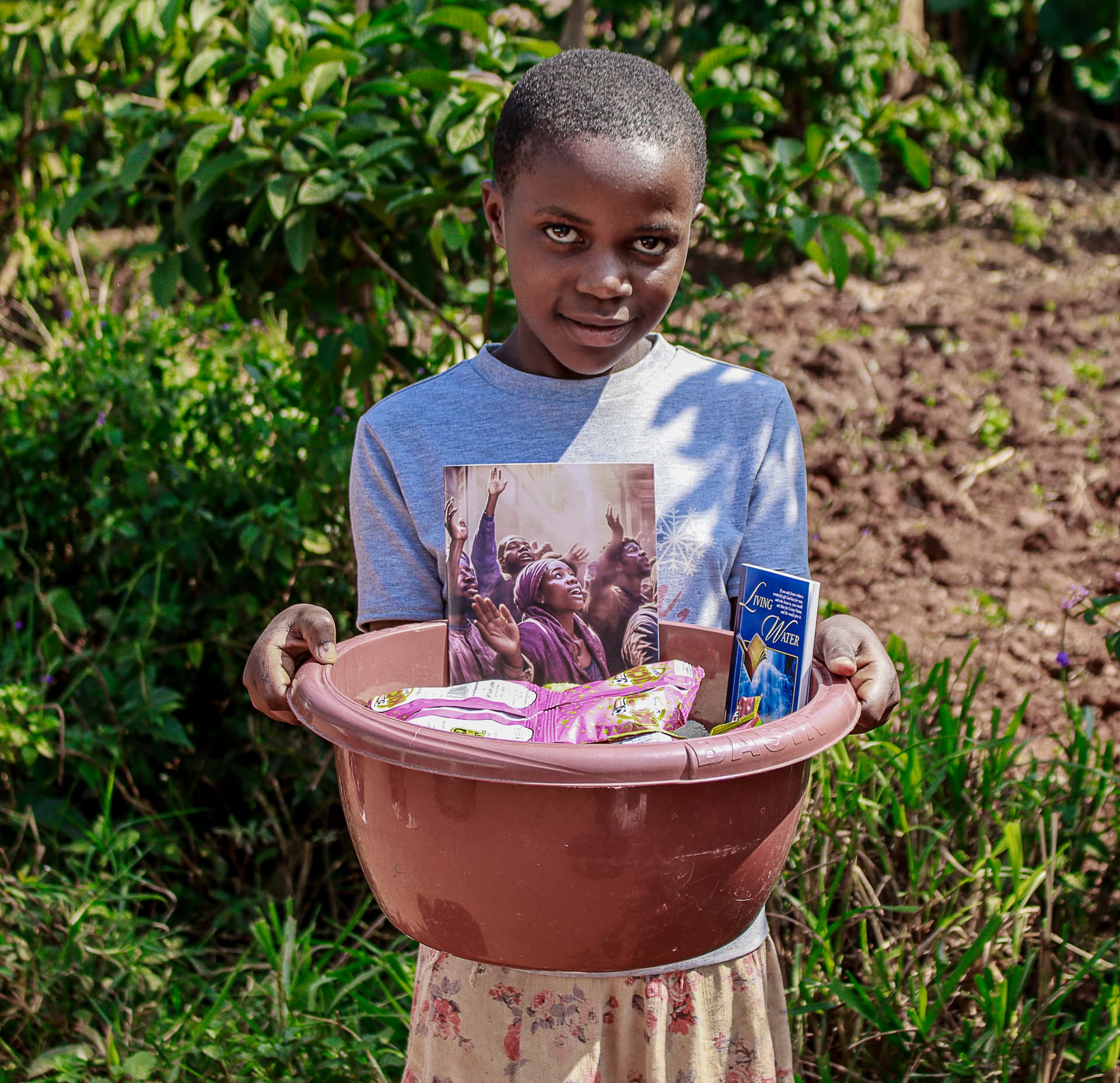 A boy stands outdoors holding a reddish basin topped with a gospel tract and food packets.