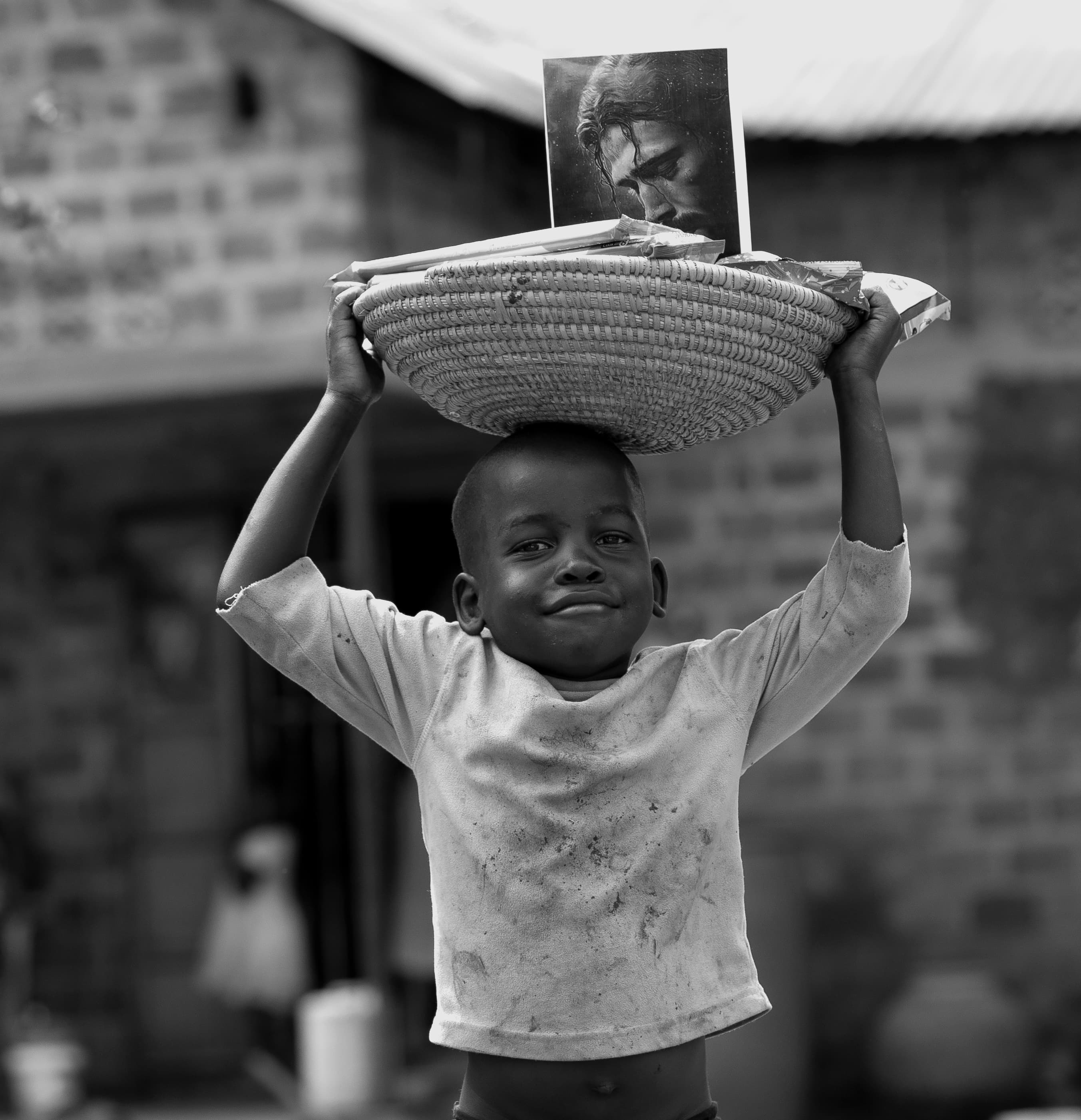 Black-and-white photo of a boy carrying a woven basket with a photo card on his head outside a brick building.