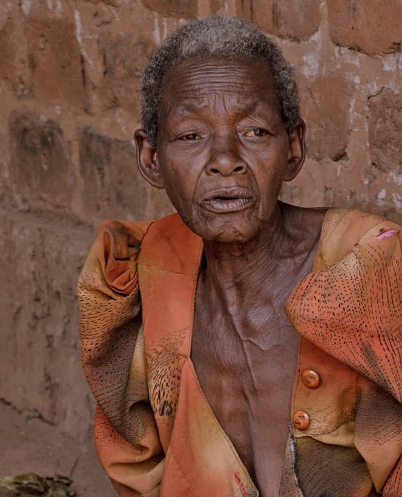 Close-up portrait of an elderly woman with a weathered face, standing against a red brick wall.