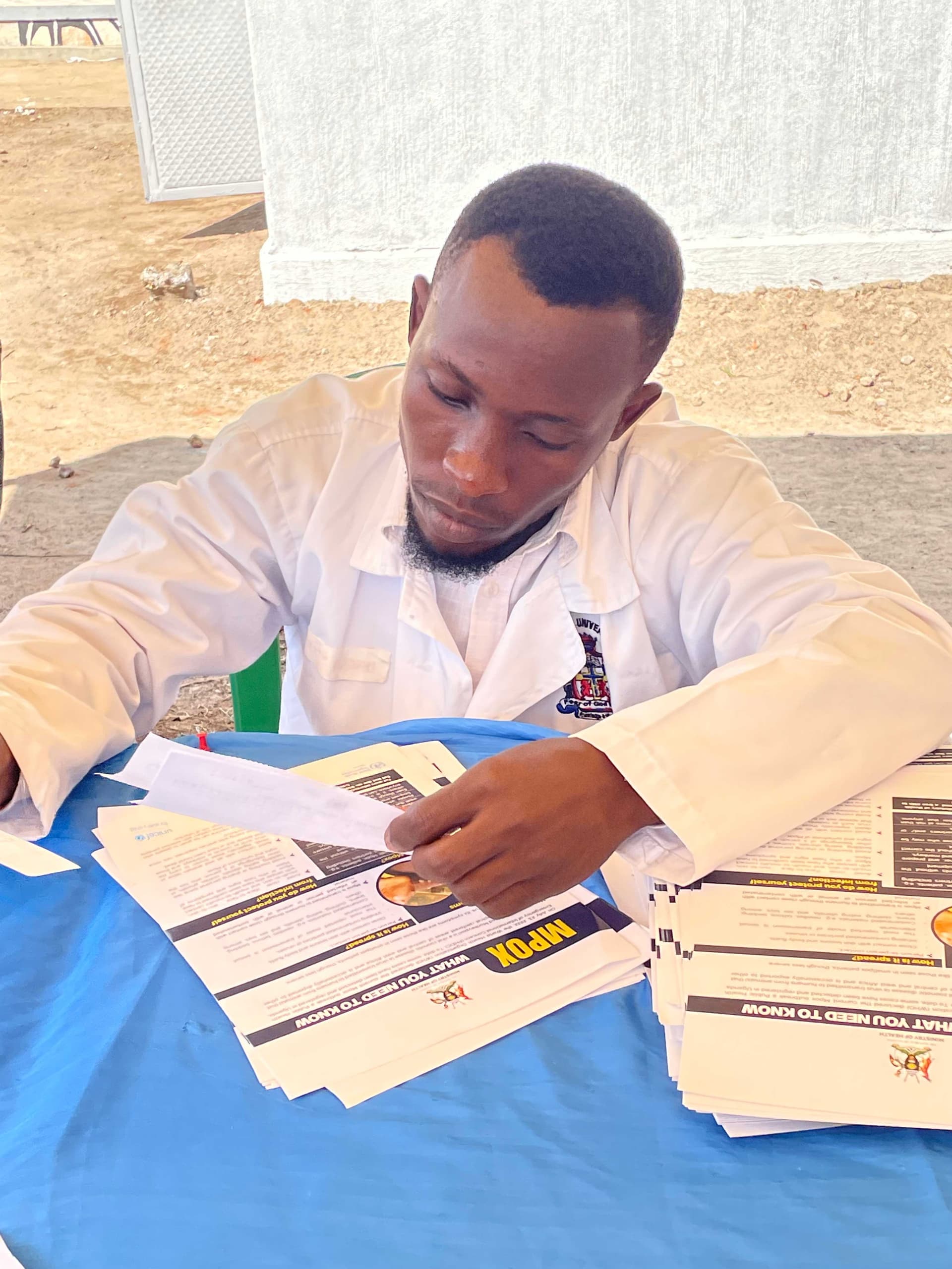 A man in a white medical coat sits at a table outdoors sorting paperwork and forms during the Miracle Medical Centre opening.