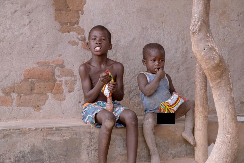 Two children sit against a wall holding packaged food items.