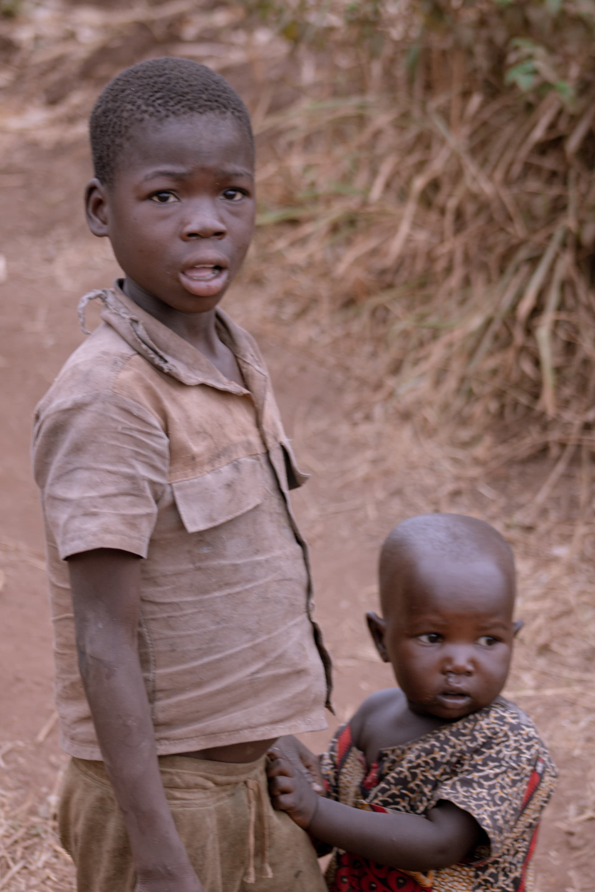 An older boy holds a younger sibling on a dirt path near a wooded area in Isunga Village.